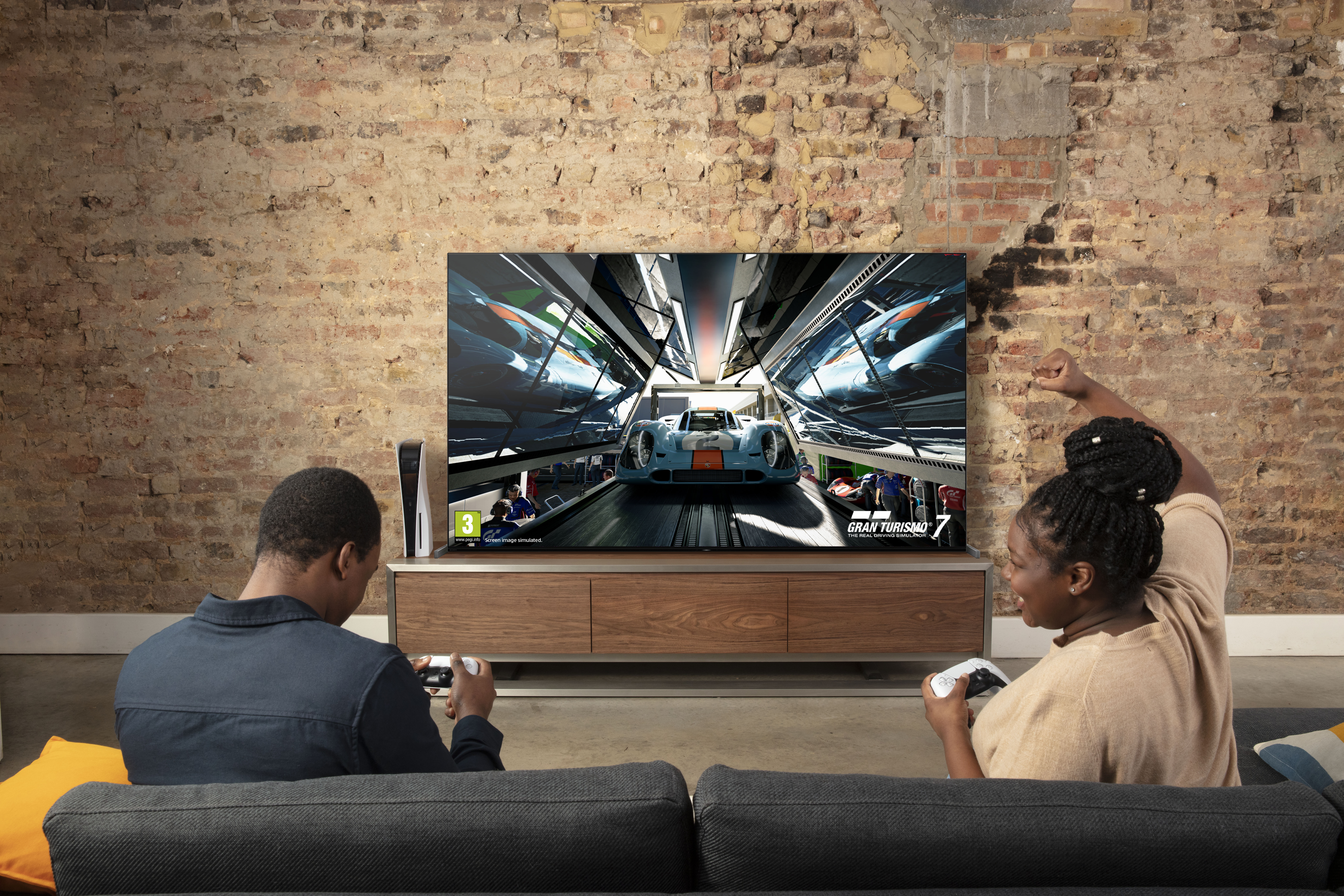 Zwei Personen spielen ein Rennspiel auf einem großen Fernseher. Eine PS5 ist neben dem Fernseher zu sehen.