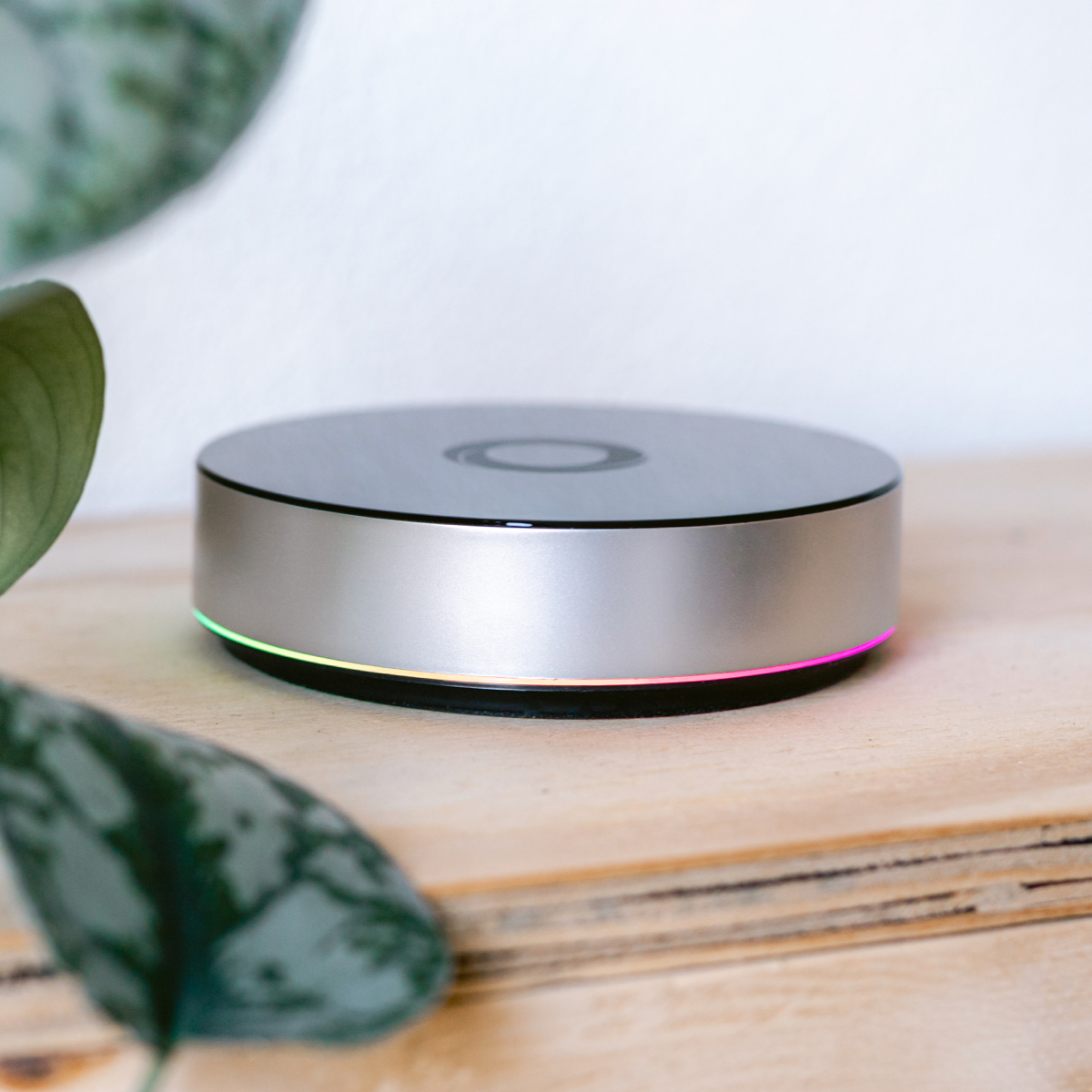Silbernes, rundes Smart-Home-Gerät mit farbigem Lichtring. Auf Holzoberfläche.