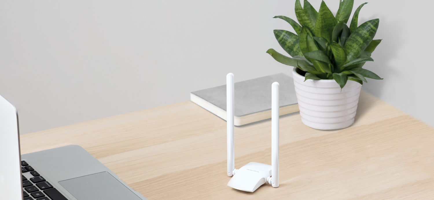 Egy router két antennával egy fa asztalon ül egy laptop és egy cserepes növény mellett.