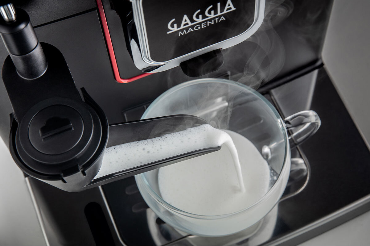 Egy fekete Gaggia Magenta eszpresszó gép. Habosított tejet öntenek egy üvegpohárba, gőz emelkedik.
