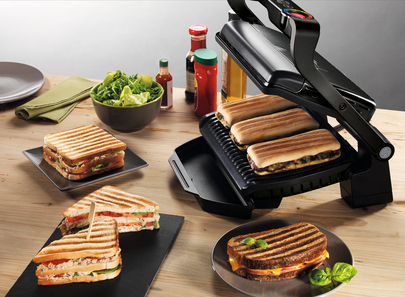Un grill à sandwich avec plusieurs sandwichs cuits et ingrédients sur une table en bois.