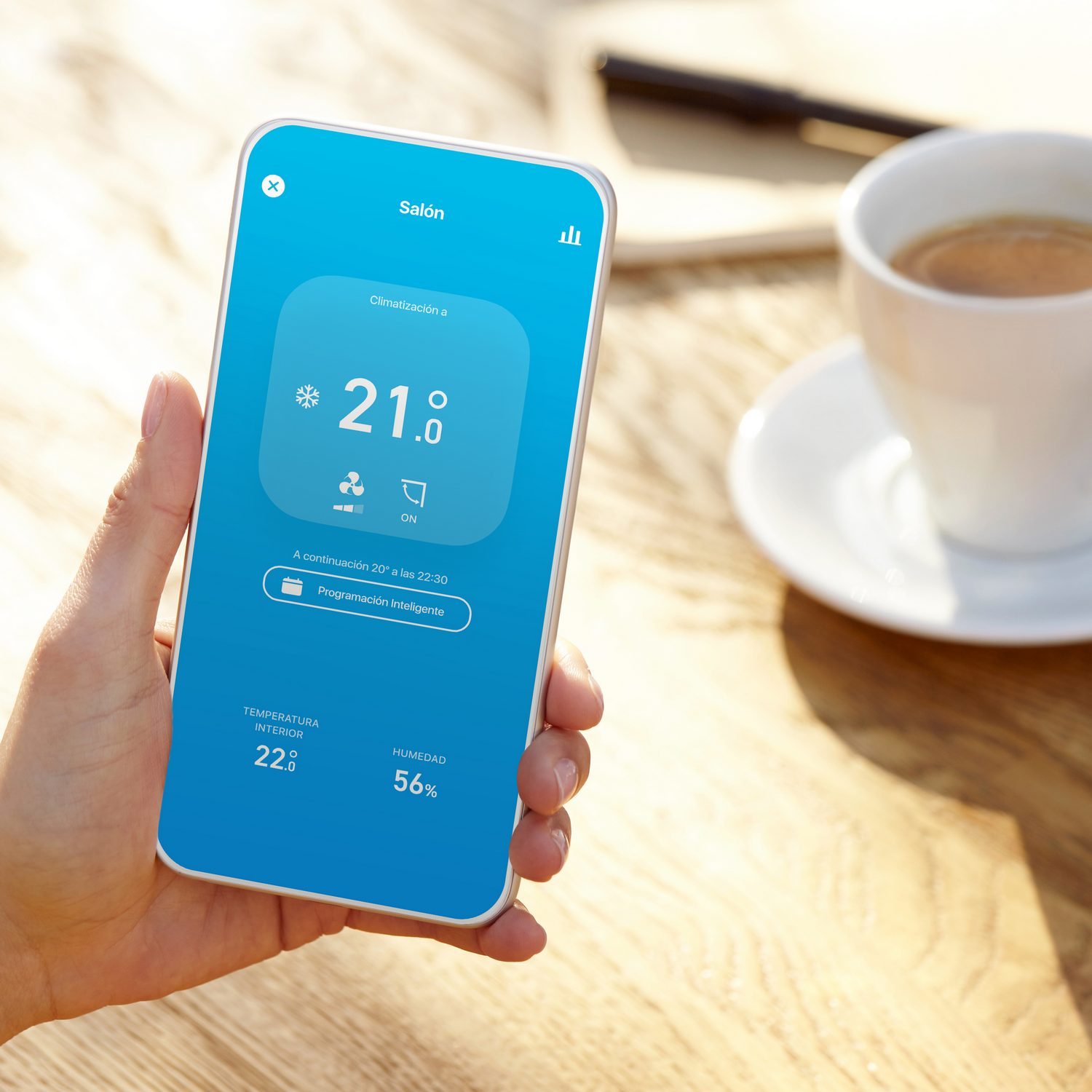 Mano sosteniendo un teléfono con app de casa inteligente, taza de café y fondo borroso.