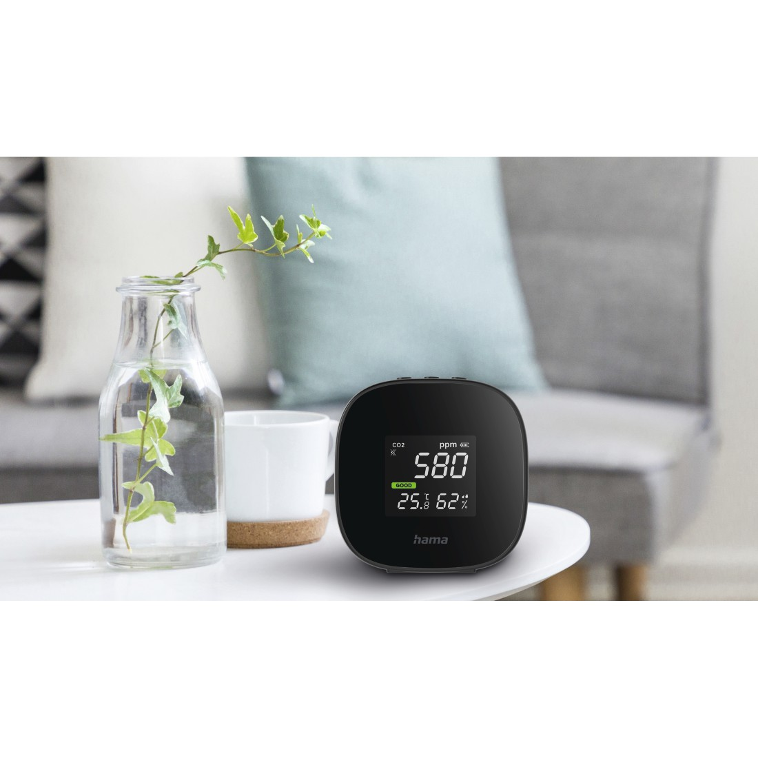 Ein schwarzer CO2-Monitor mit Anzeige der Messwerte. Eine Vase und eine Tasse stehen auf dem Tisch.