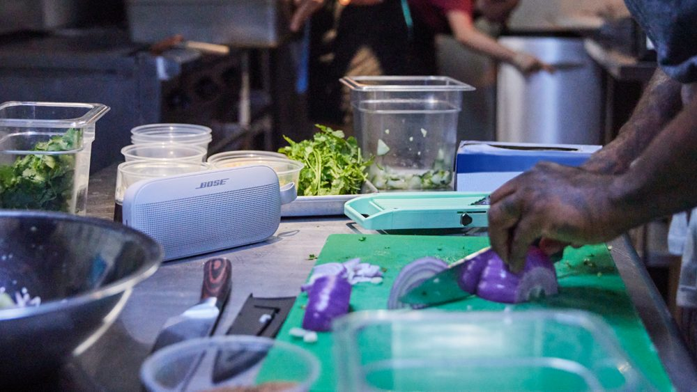 Un haut-parleur Bose sur un comptoir, avec une personne coupant des légumes en arrière-plan.