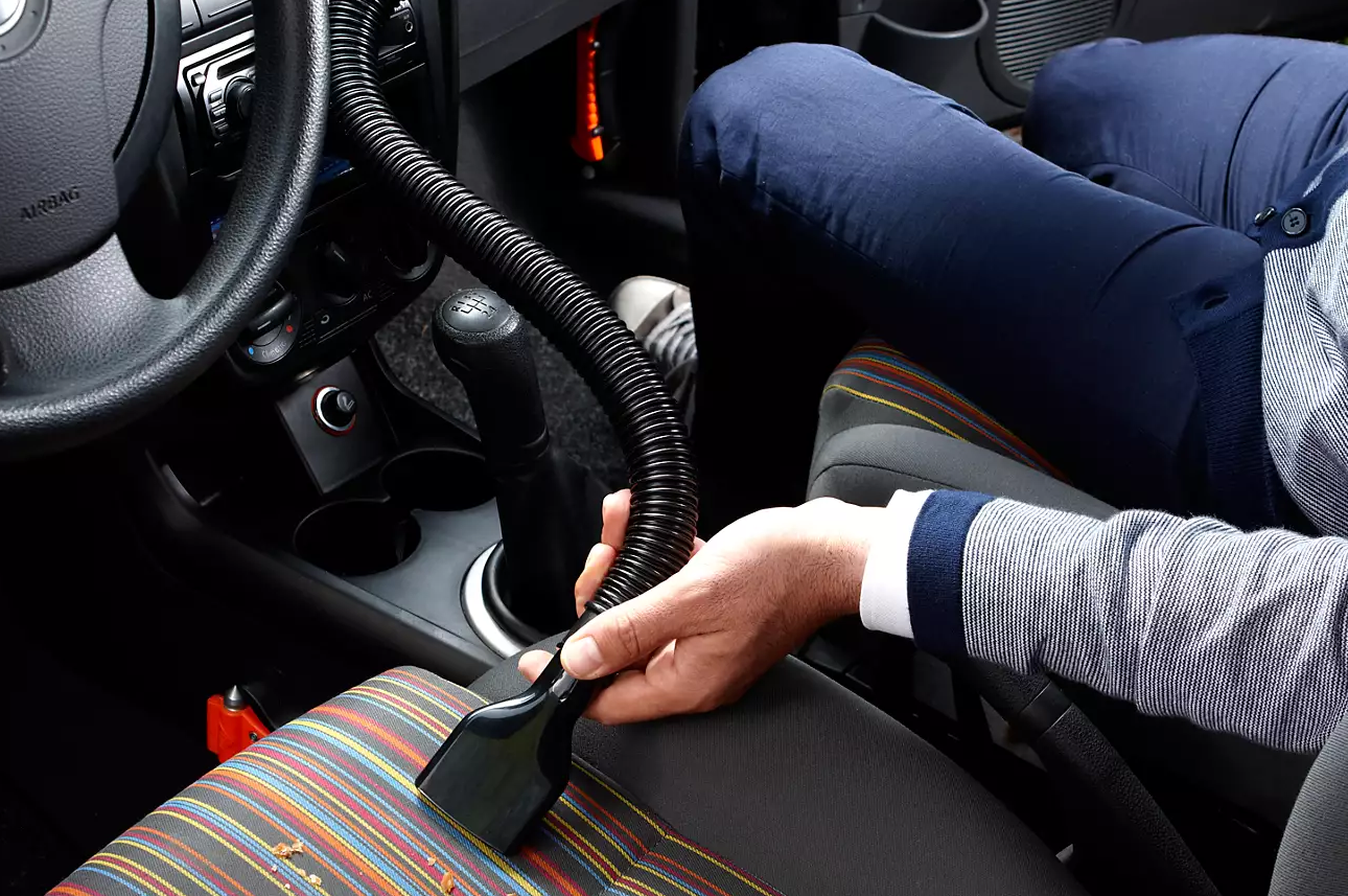 Personne nettoyant un siège de voiture avec un aspirateur. L'intérieur de la voiture est visible.