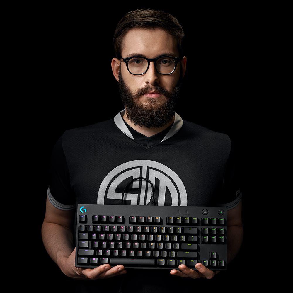 Man in glasses holding a black keyboard. TSM logo on shirt. Black background.