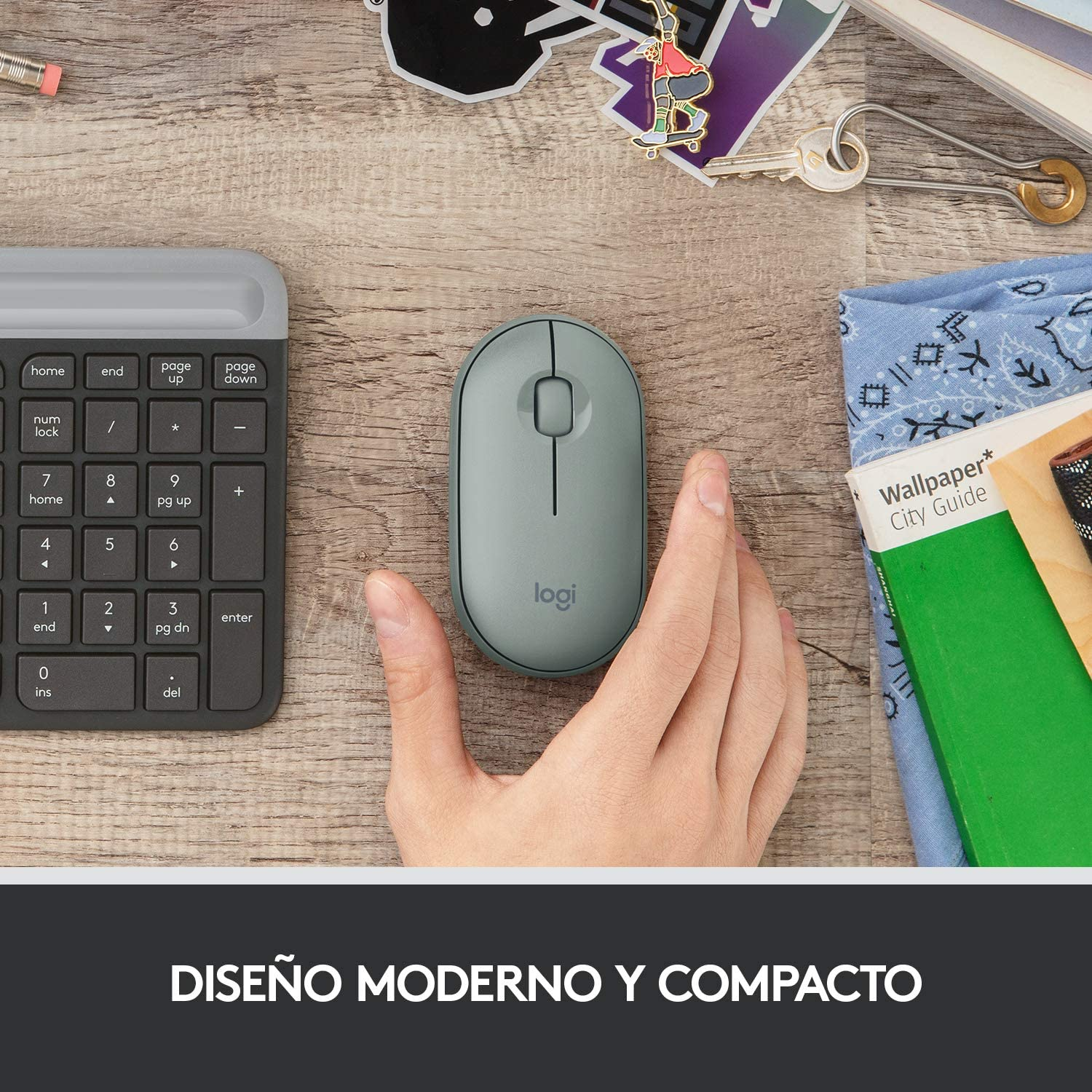 Mano sosteniendo un ratón logi gris junto a un teclado y otros objetos de escritorio.