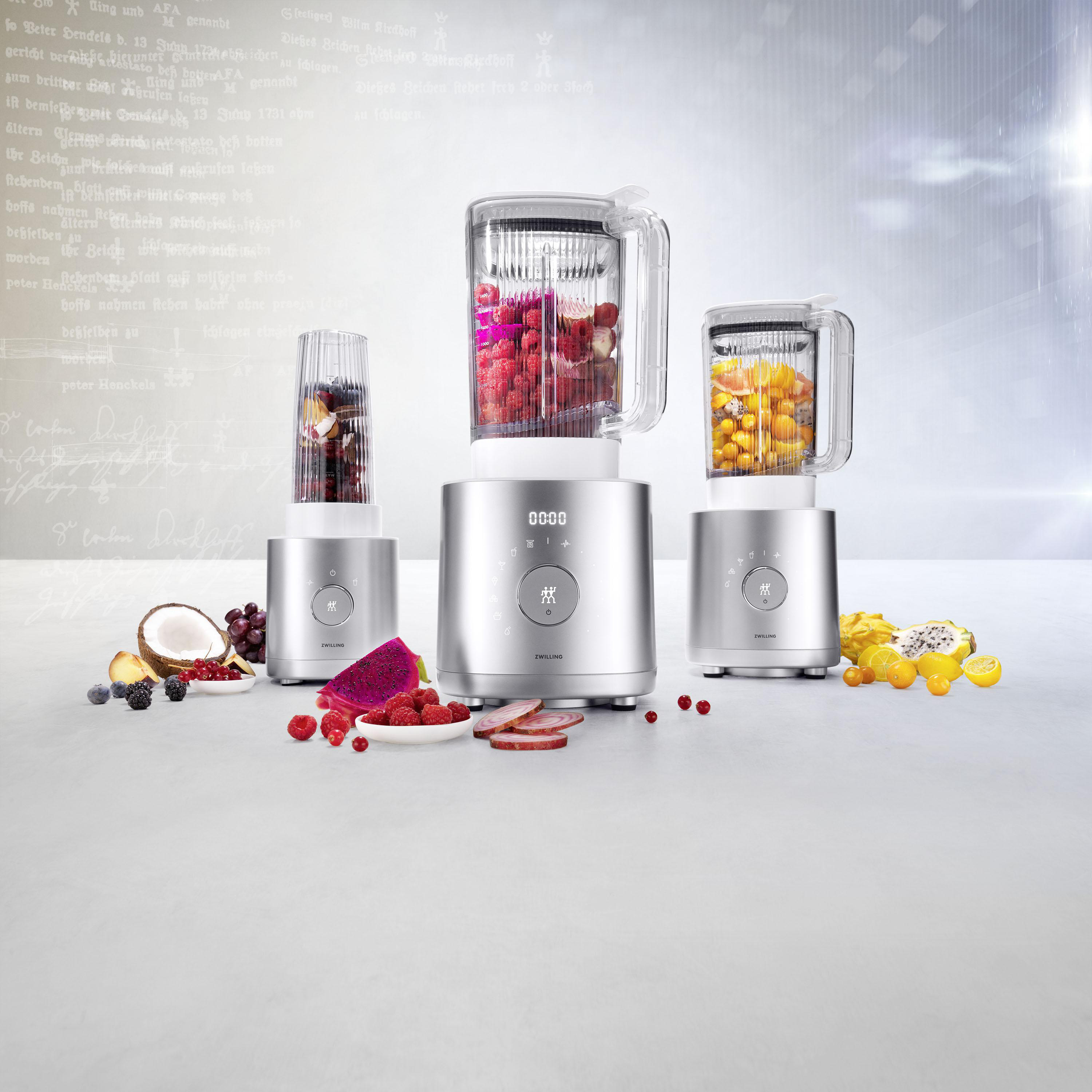 Drei silberne Mixer mit verschiedenen Früchten. Kokosnuss und andere Früchte um die Mixer herum.