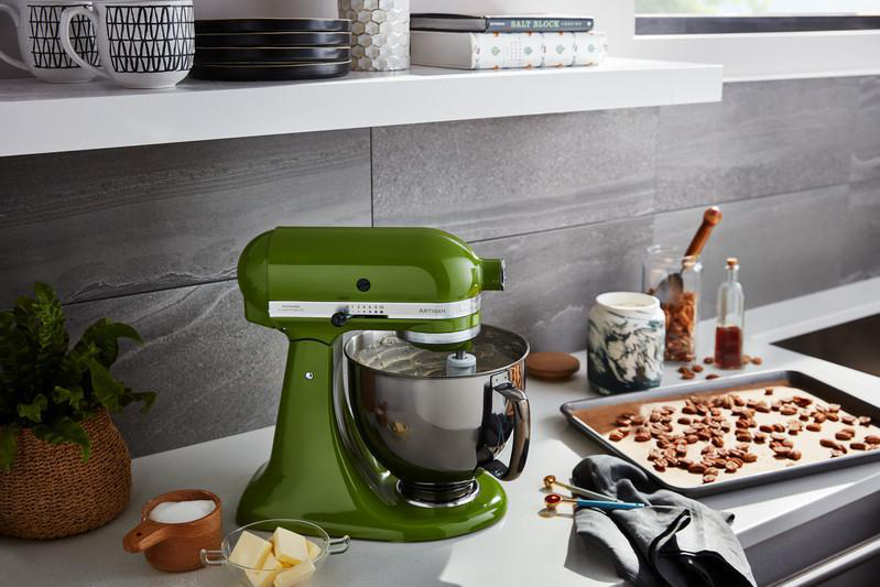 Ein grüner KitchenAid-Küchenmixer mit einer silbernen Schüssel auf einer weißen Arbeitsplatte, mit Backutensilien.