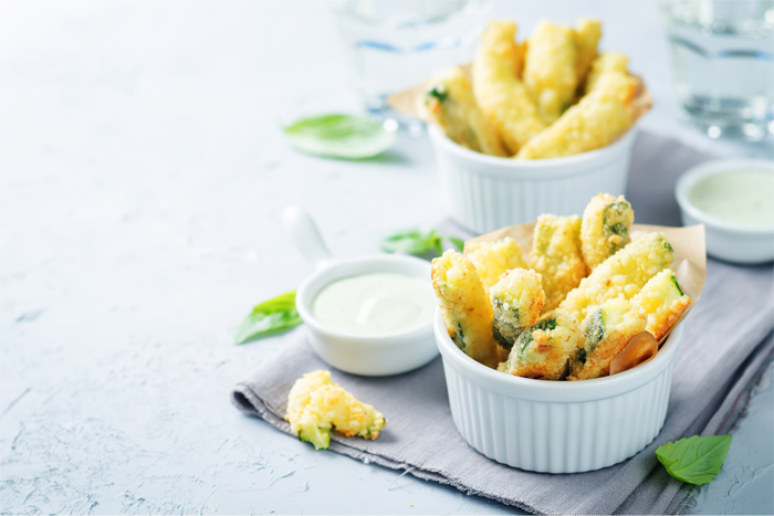 Gefrituurde courgettesticks in papieren bekers, met dipsaus en basilicumblaadjes. Heldere witte achtergrond.