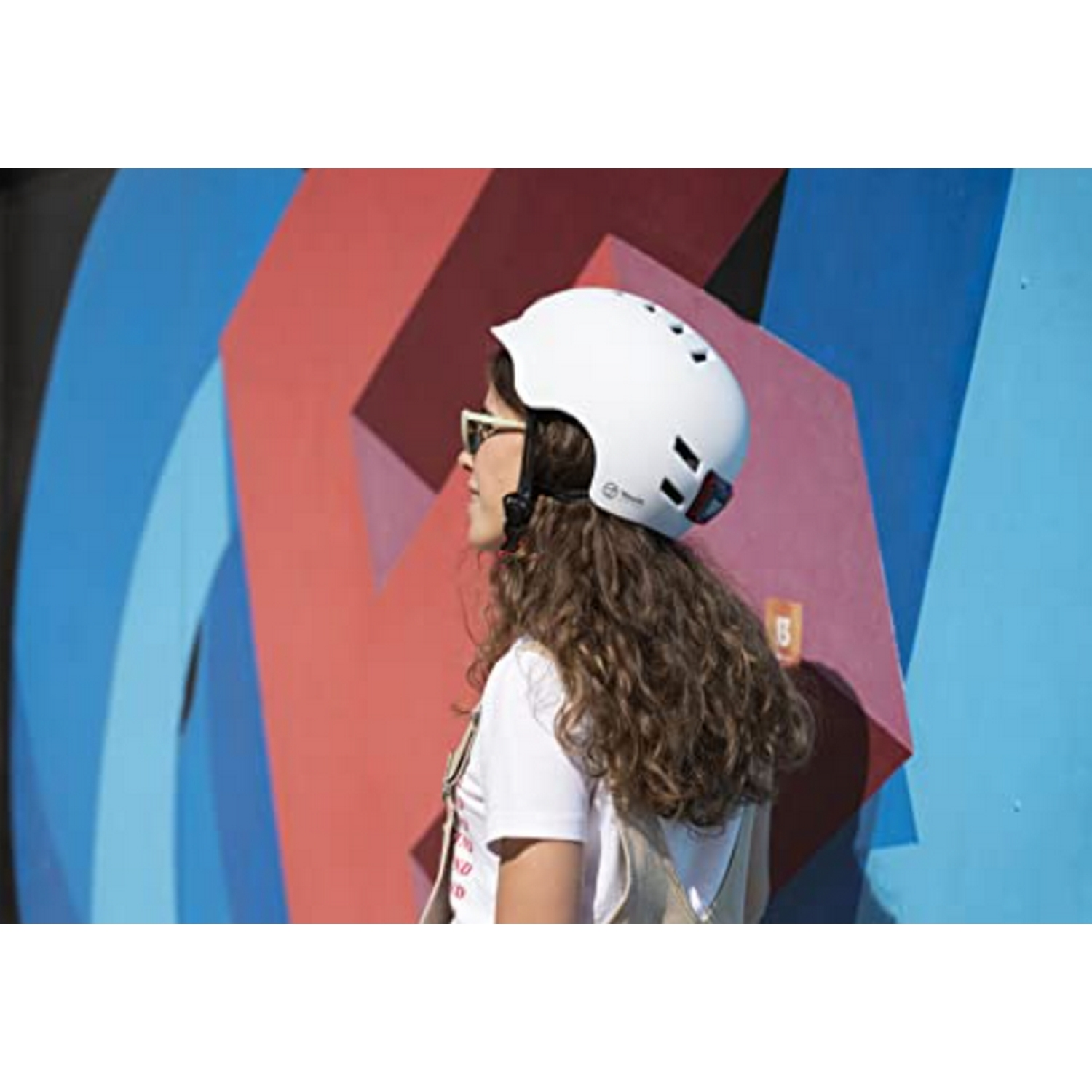 Mujer con casco blanco y gafas de sol contra mural geométrico colorido.