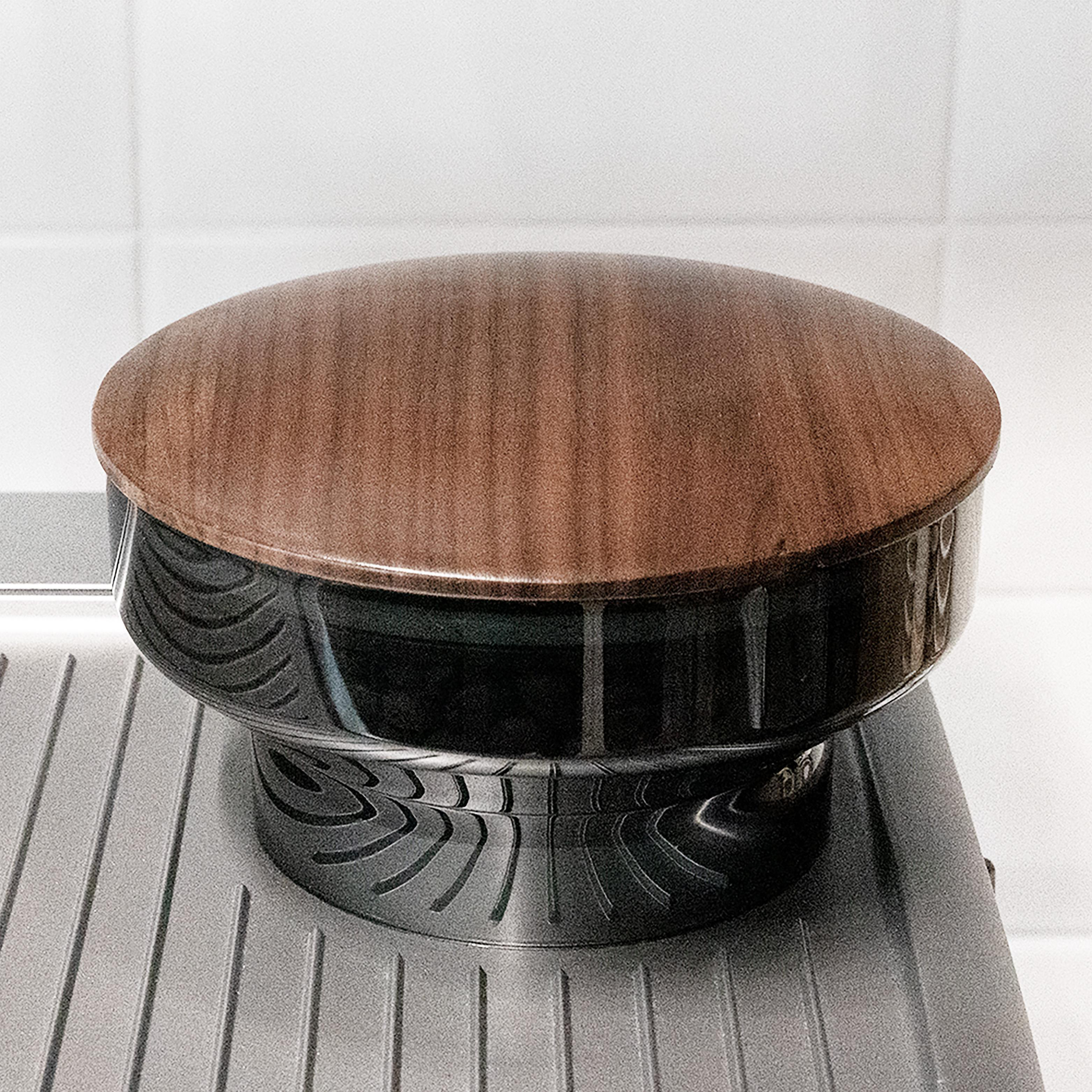 Ein brauner Holzdeckel sitzt auf einem schwarzen, abgerundeten Sockel auf einer grauen Oberfläche, vor einem weißen Fliesenhintergrund.