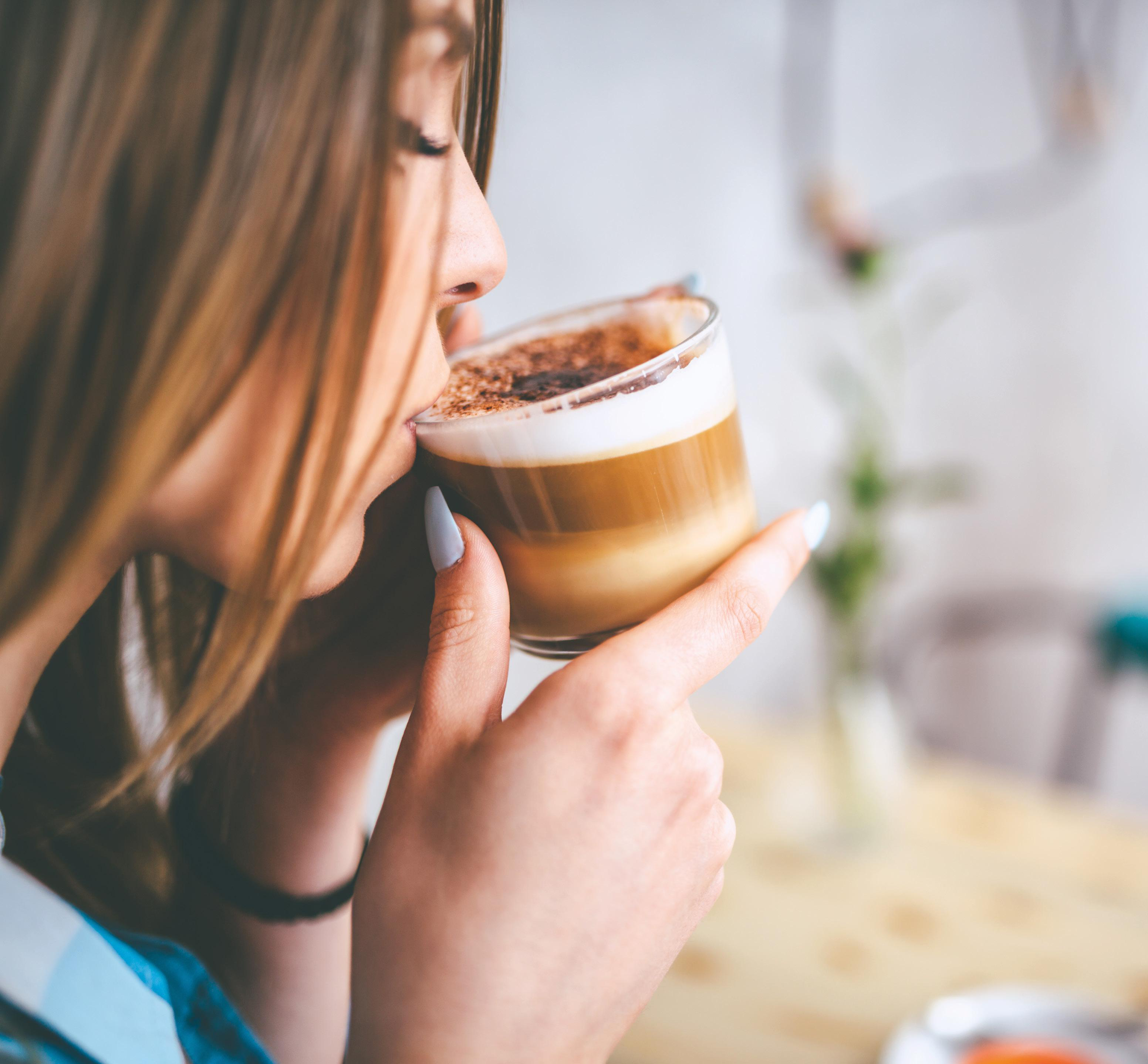 Frau trinkt Kaffee aus einem Glas. Schaumiger Belag und hellbraune Flüssigkeit. Verschwommener Hintergrund.