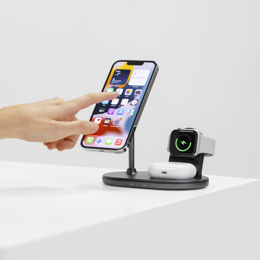 Eine Hand berührt ein Telefon auf einem Ständer. Eine Apple Watch und Ohrhörer befinden sich neben dem Telefon.