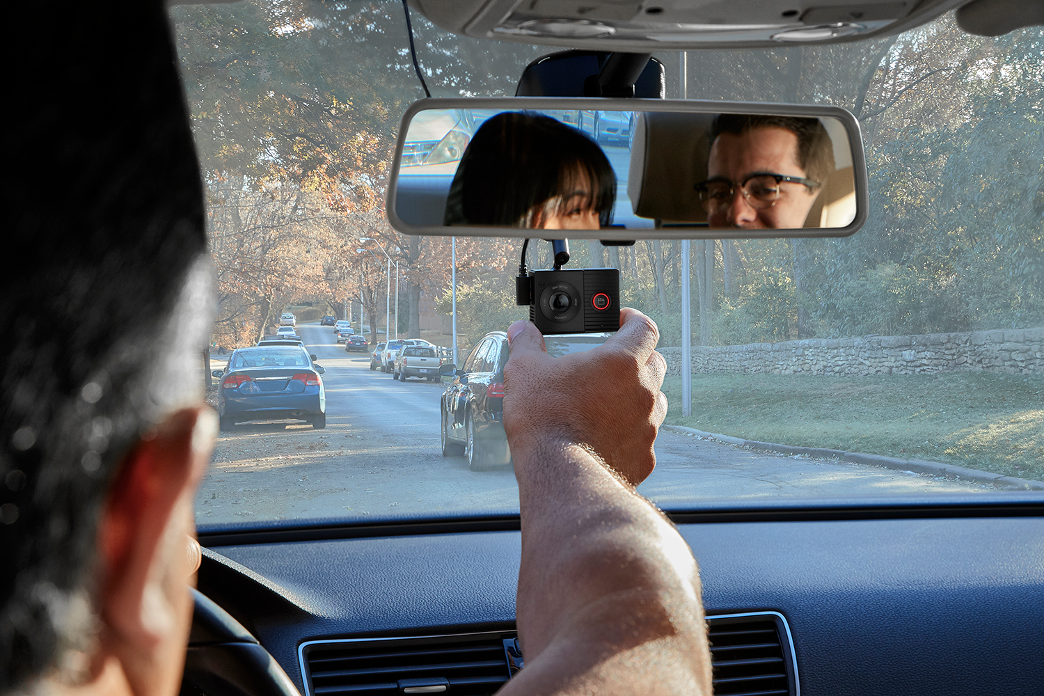 Person in a car holding a dashcam, reflecting in the rearview mirror with two people.