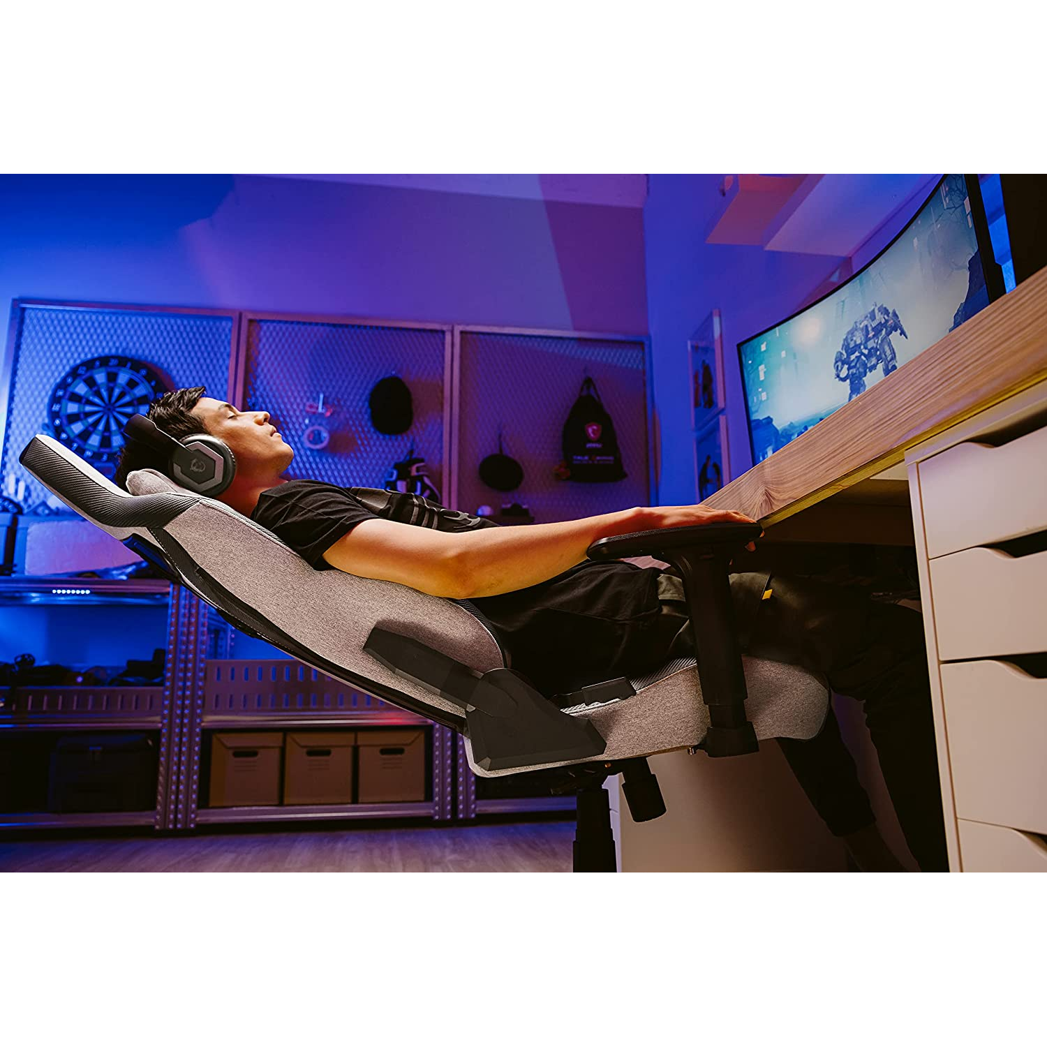 Un hombre reclinado en una silla gaming, con auriculares, con un monitor curvo al fondo.