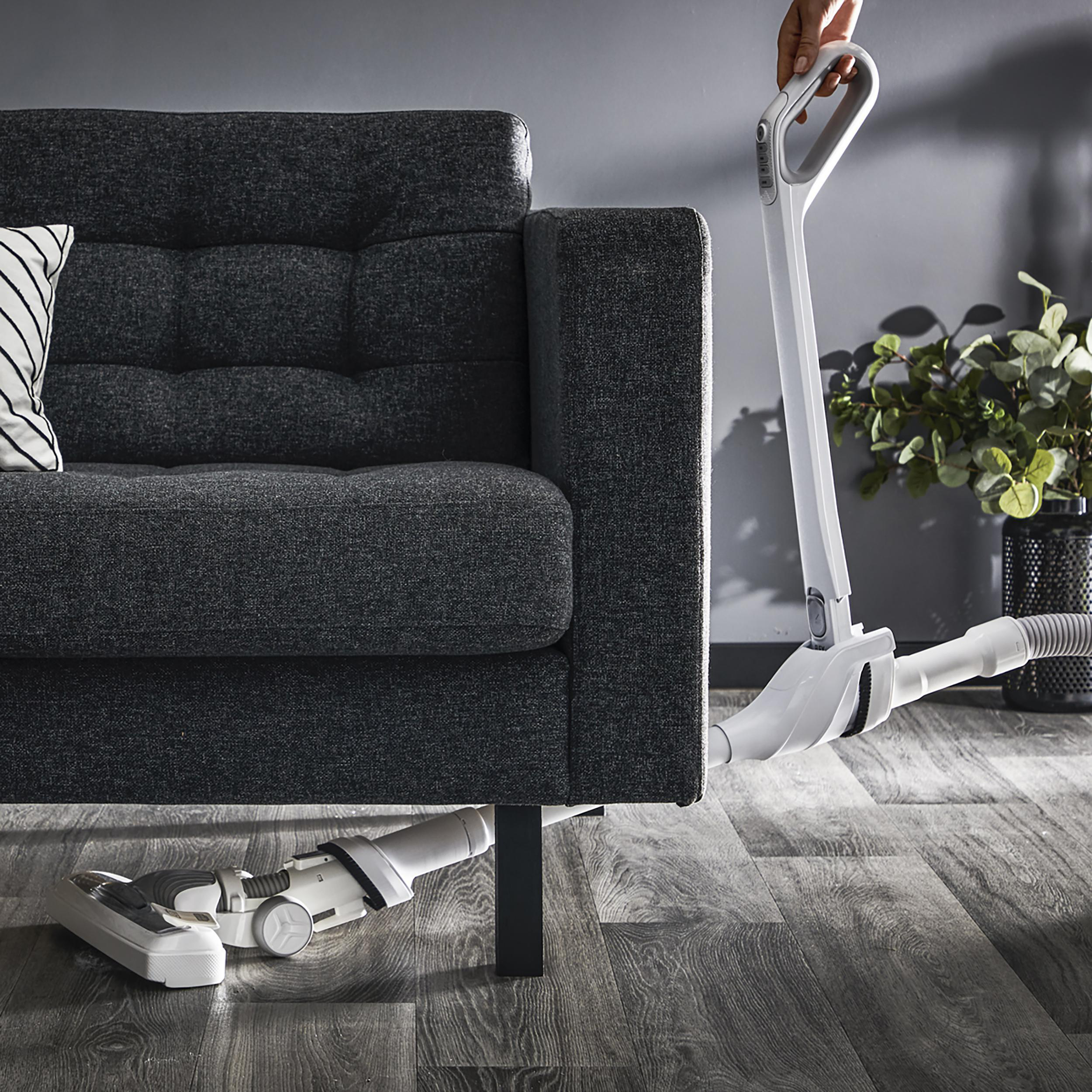 Staubsauger reinigt unter einem grauen Sofa. Der Staubsauger ist weiß.