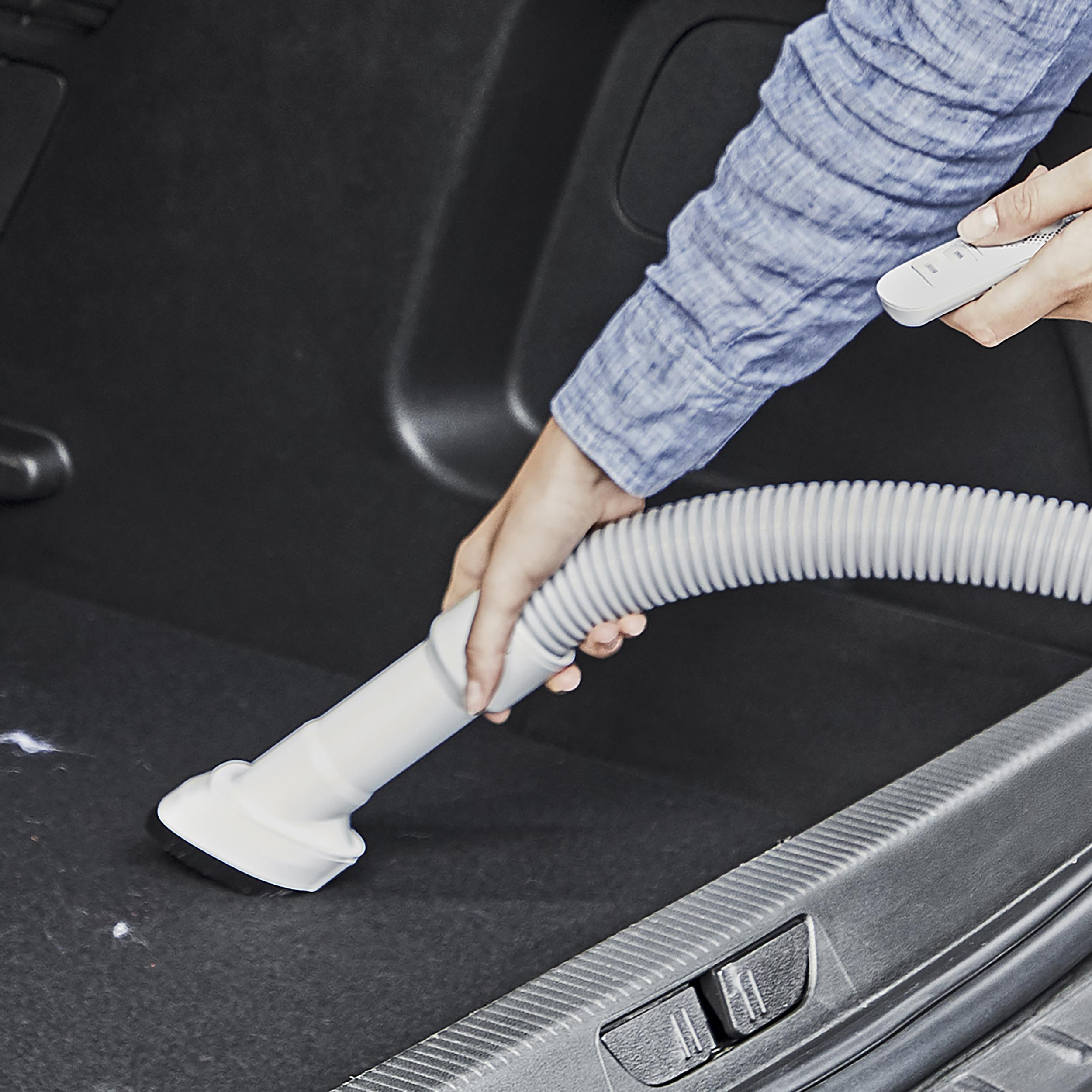 Eine Person, die den Kofferraum eines Autos saugt. Der Staubsauger hat einen weißen Schlauch und einen Bürstenkopf.