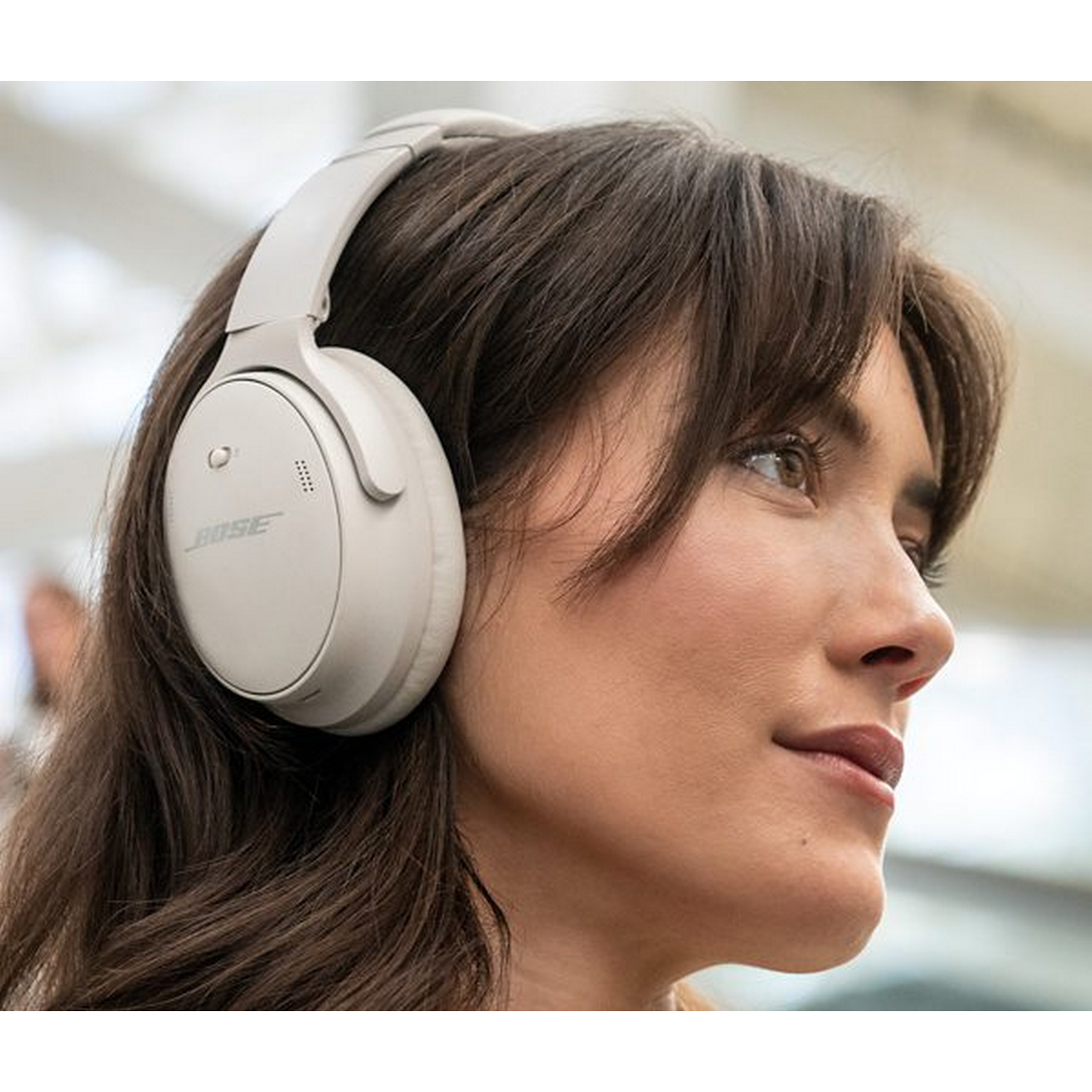 Mujer con auriculares blancos, mirando a la derecha. Cabello castaño, fondo borroso.