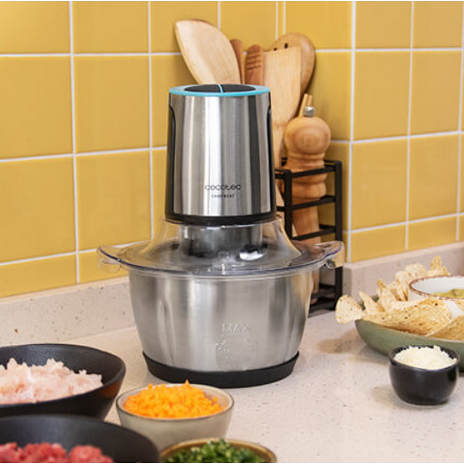 Un picador de alimentos de acero inoxidable con cuencos de ingredientes y fondo de azulejos amarillos.