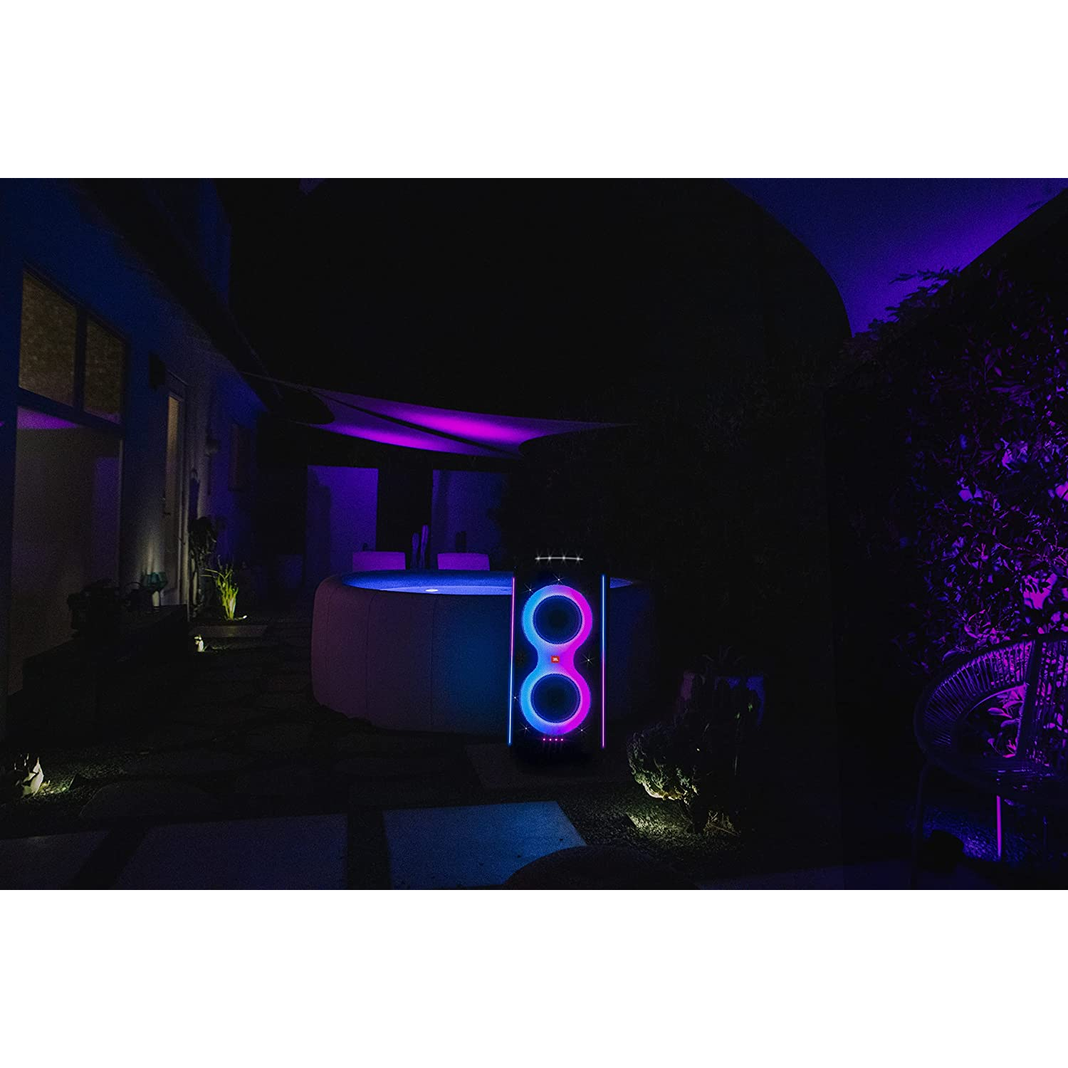 Patio trasero por la noche con un altavoz que brilla en azul y rosa cerca de una bañera de hidromasaje y plantas.