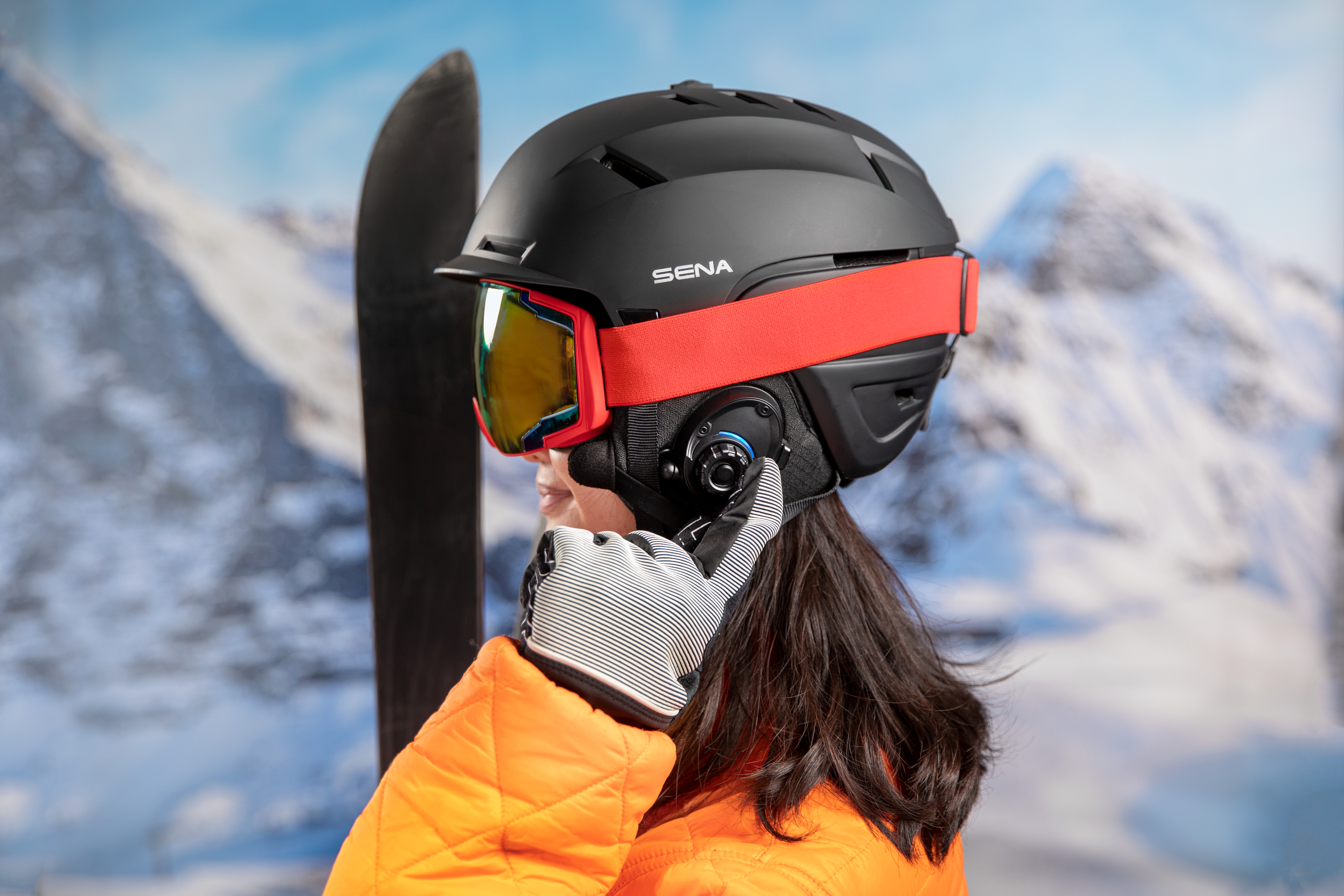 Eine Person trägt einen schwarzen Skihelm mit Brille und rotem Band. Ein Handschuh berührt den Helm.