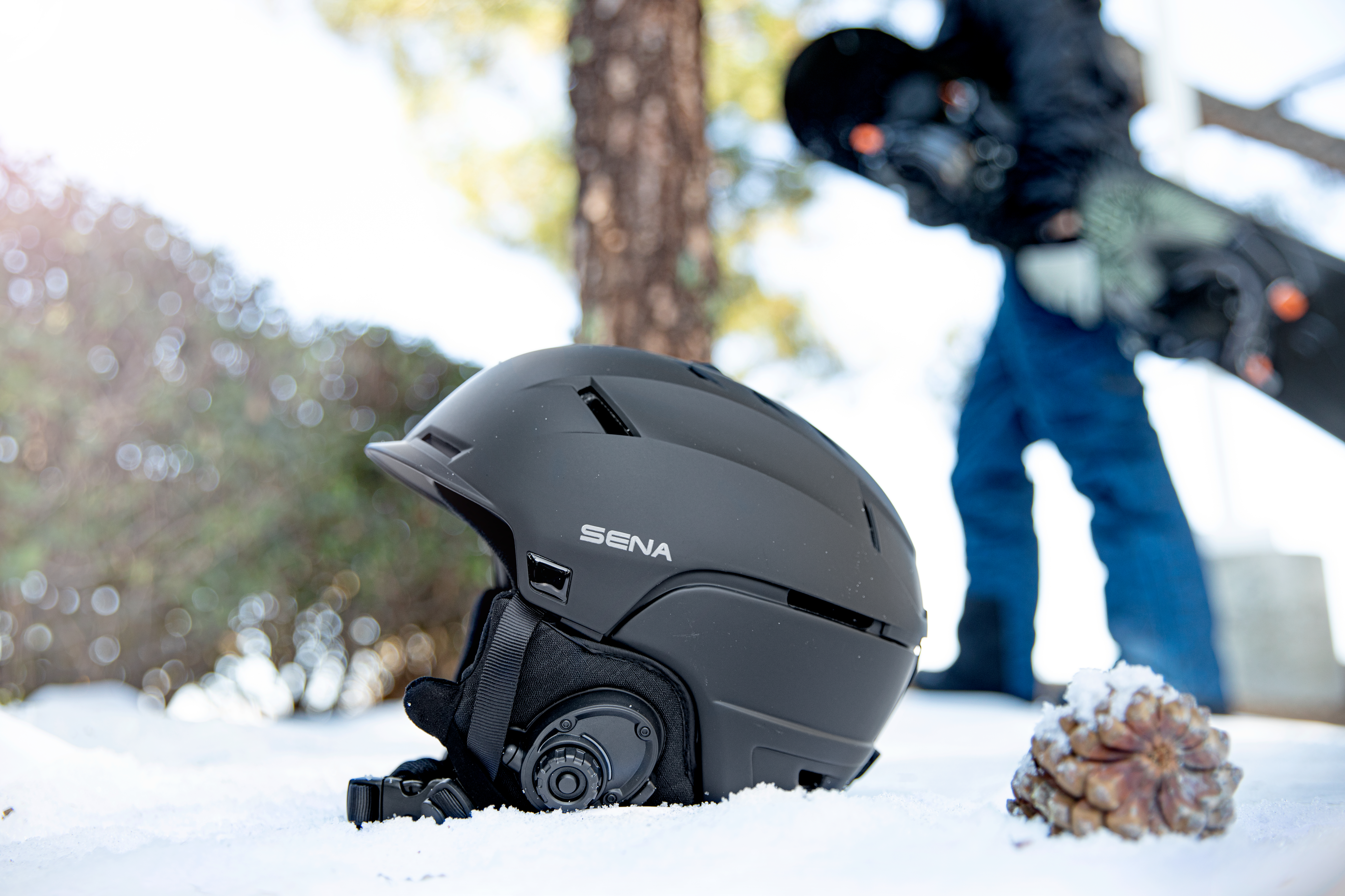 Ein schwarzer Helm liegt im Schnee. Eine Person mit einem Snowboard befindet sich hinter dem Helm.