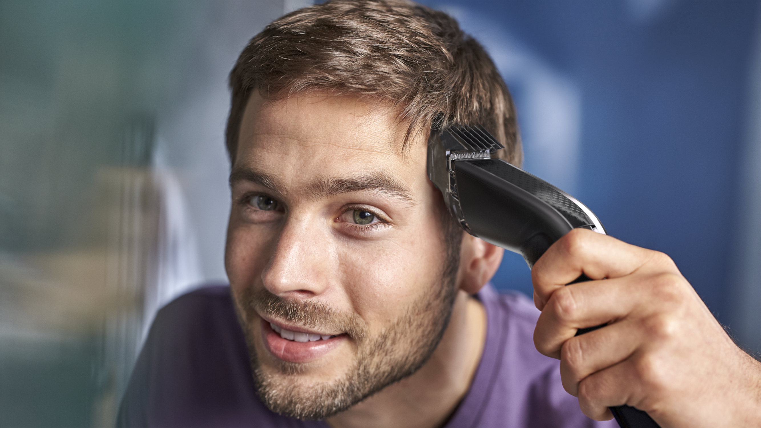 Man using a hair clipper, smiling. Blue background.
