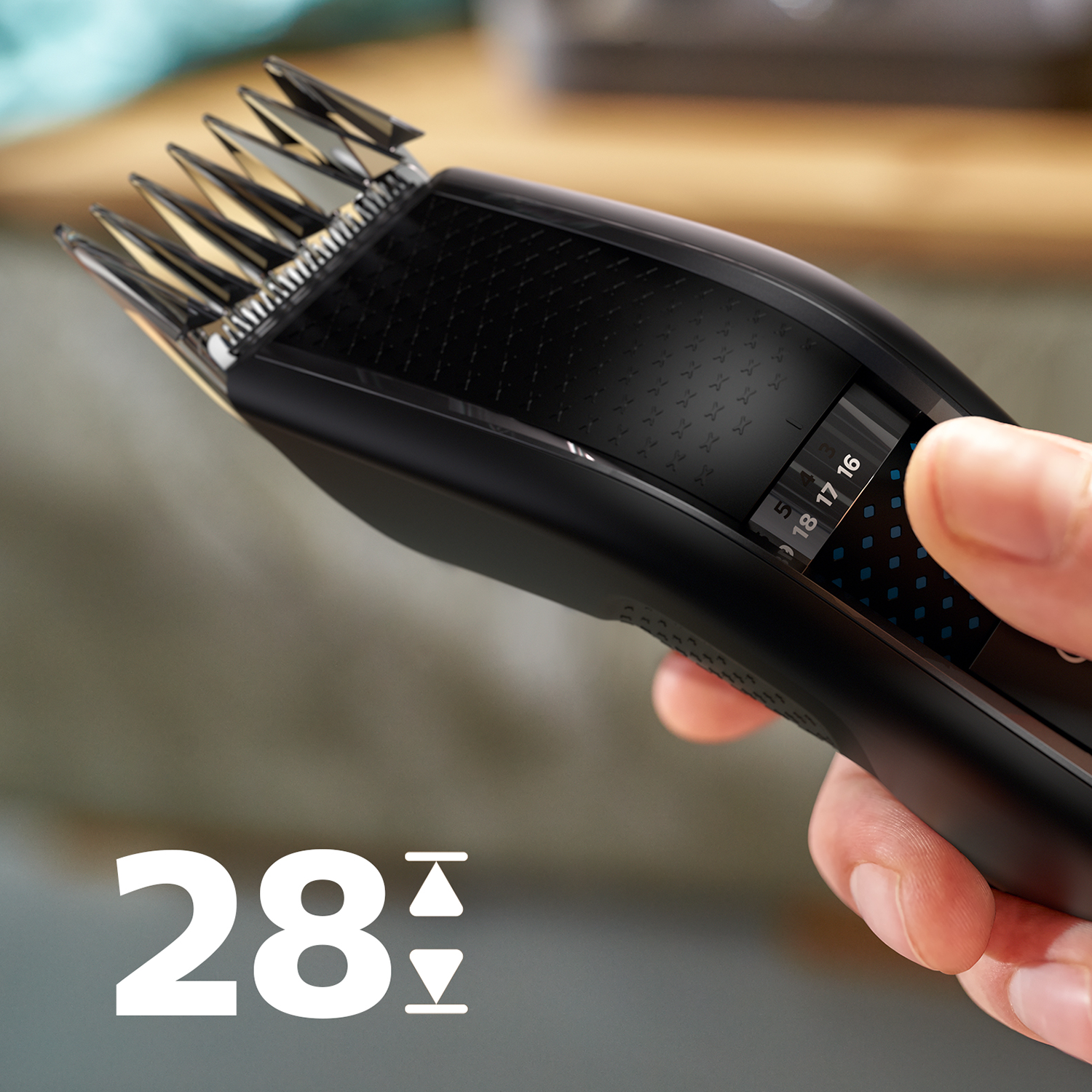 Black hair clipper, showing settings for hair length. Hand holding the clipper. Blurred background.