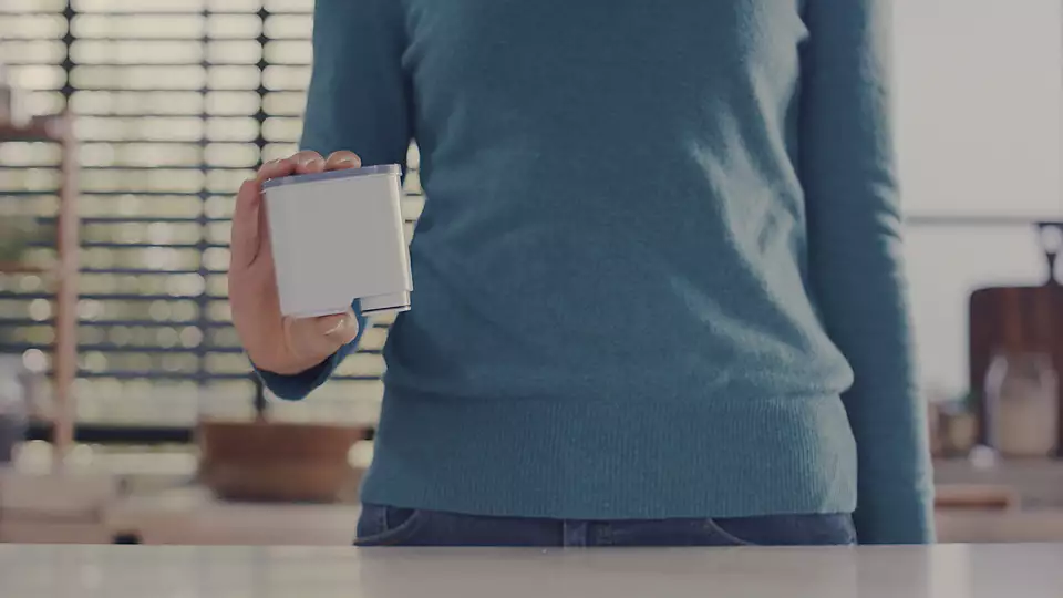 A person's hand is holding a white, rectangular cartridge. The kitchen is in the background.
