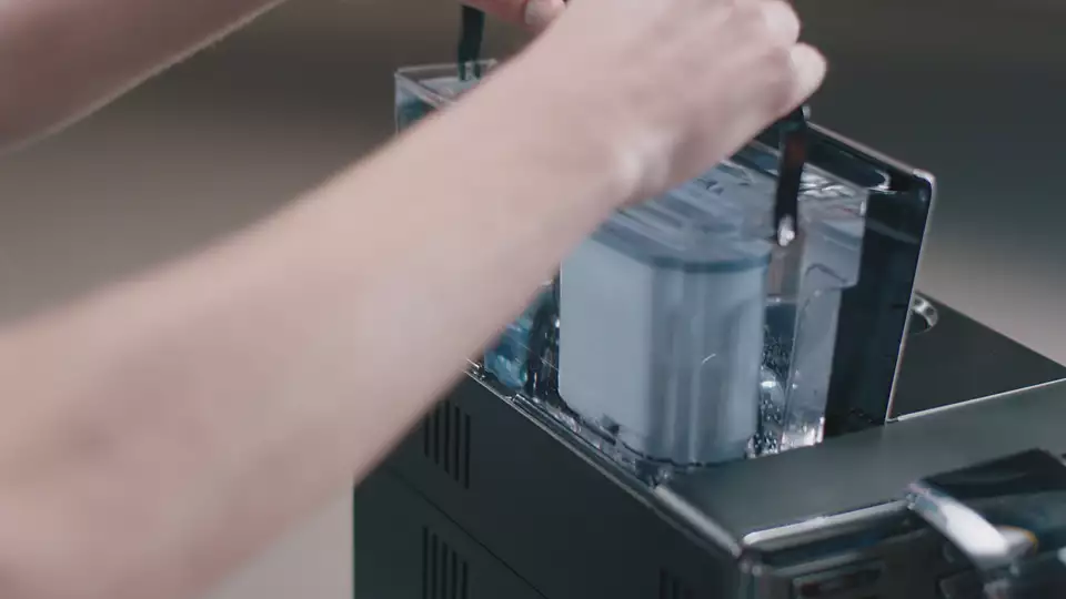 Hands insert a filter into a transparent water tank on a coffee machine.