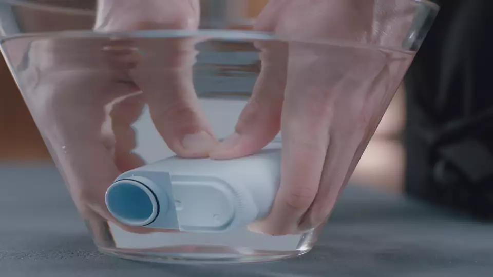Hands holding a white filter submerged in water inside a clear glass bowl, demonstrating a cleaning process.