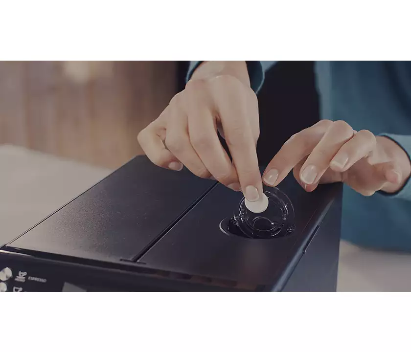Hands inserting a white disc into a dark espresso machine. The machine has the espresso and power buttons.