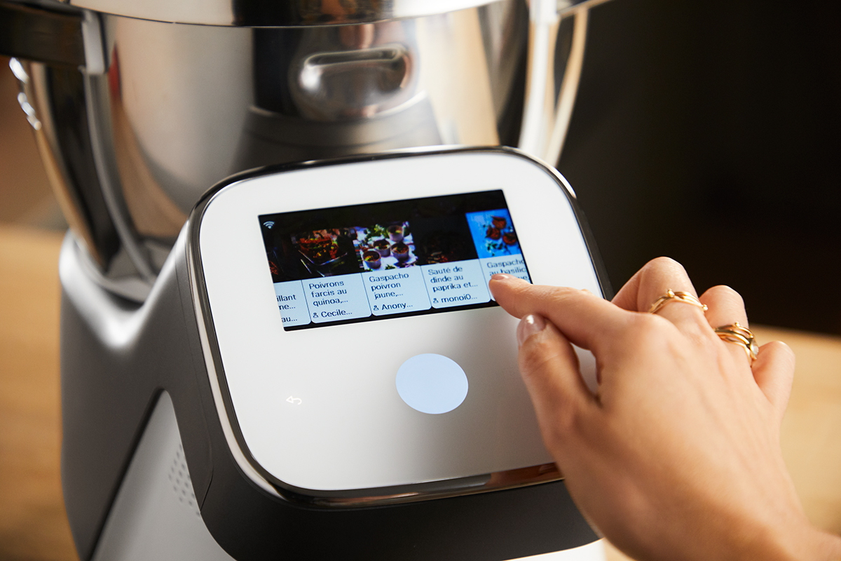 Une main touche l'écran d'un appareil de cuisine intelligent affichant un menu de recettes.