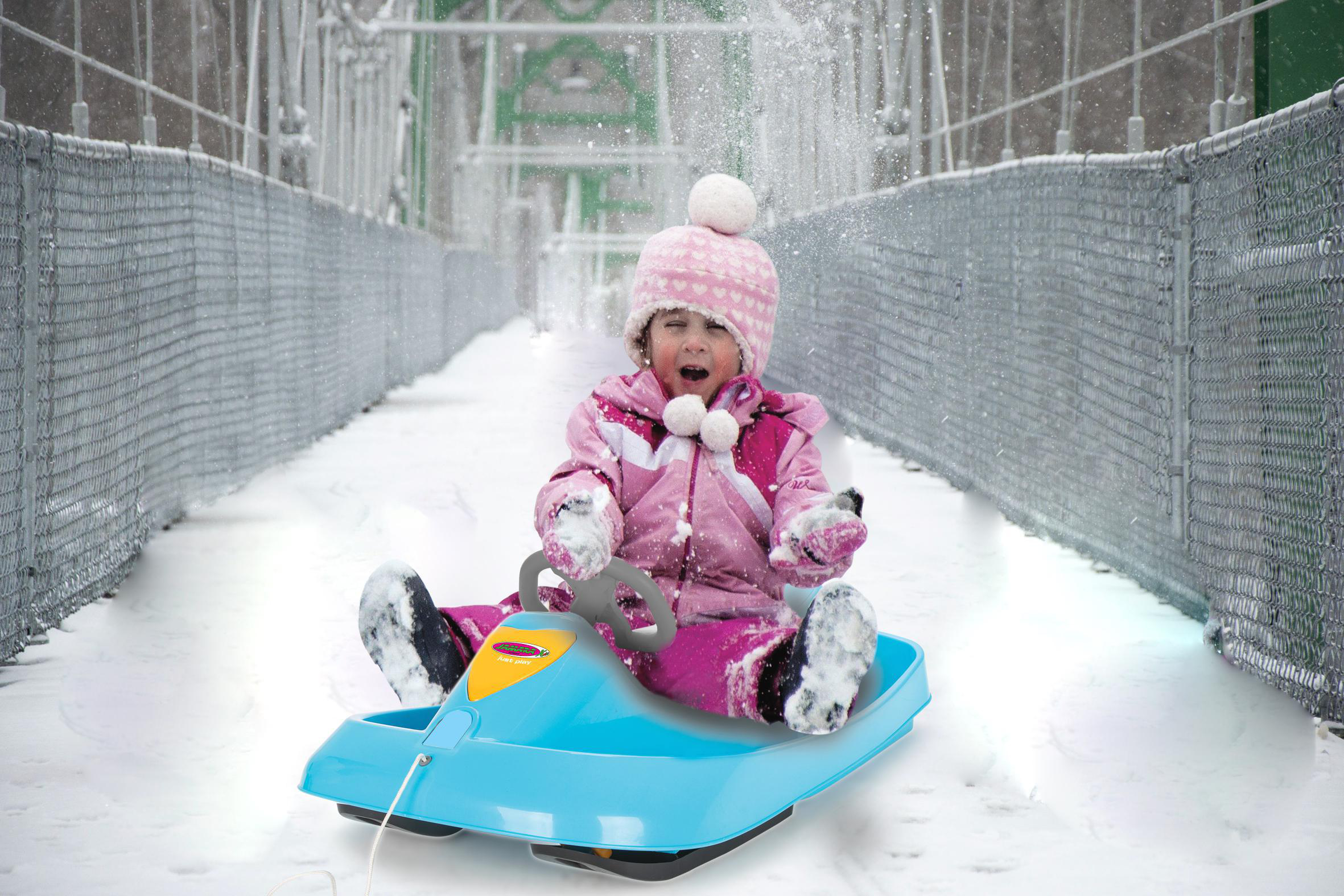 Kind schlittenfährt über schneebedeckte Brücke. Rosa Outfit, blauer Schlitten. Schnee fällt.