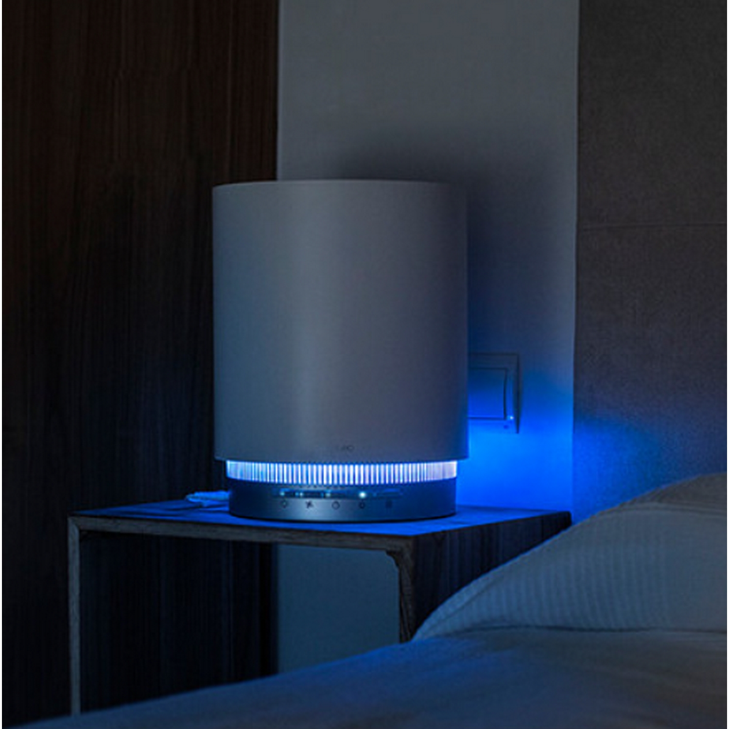 Un purificador de aire blanco brilla en azul. Está sobre una mesita de noche de madera junto a una cama con sábanas blancas. La pared es oscura.