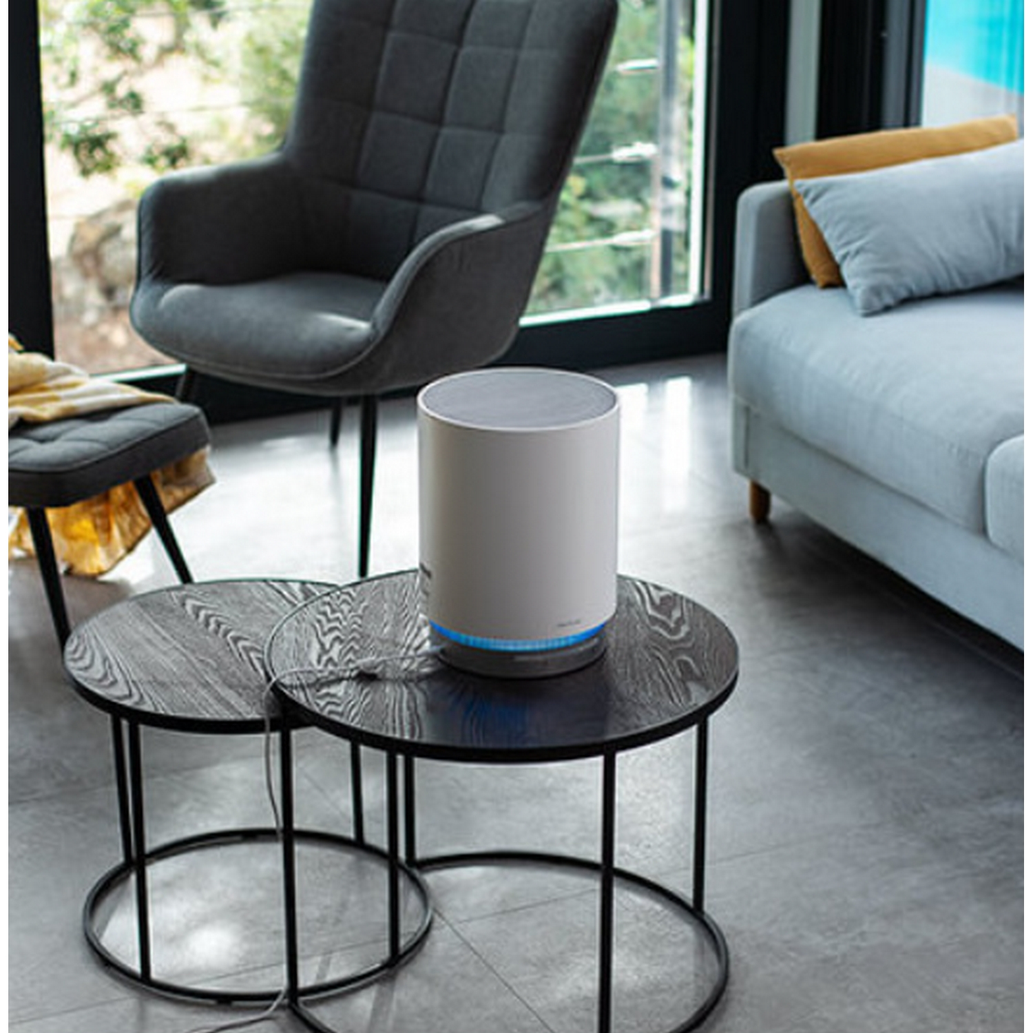 Un altavoz blanco sobre una mesa negra. Silla gris, sofá y mesita en el fondo. La luz natural entra por una ventana.