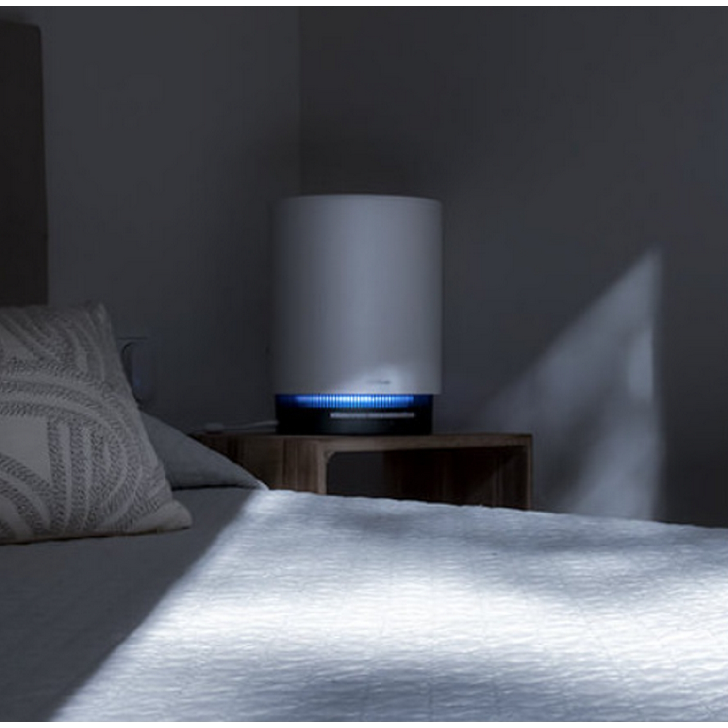 Un purificador de aire blanco con luz azul se encuentra en una mesita de noche de madera junto a una cama con ropa de cama blanca y una almohada.