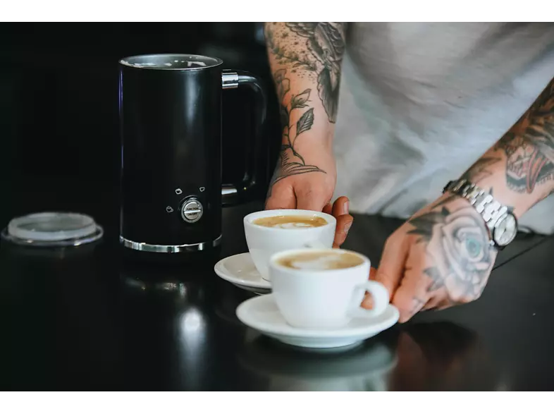 A barista serving two cappuccinos. A black milk frother is on the left, tattoos are visible.