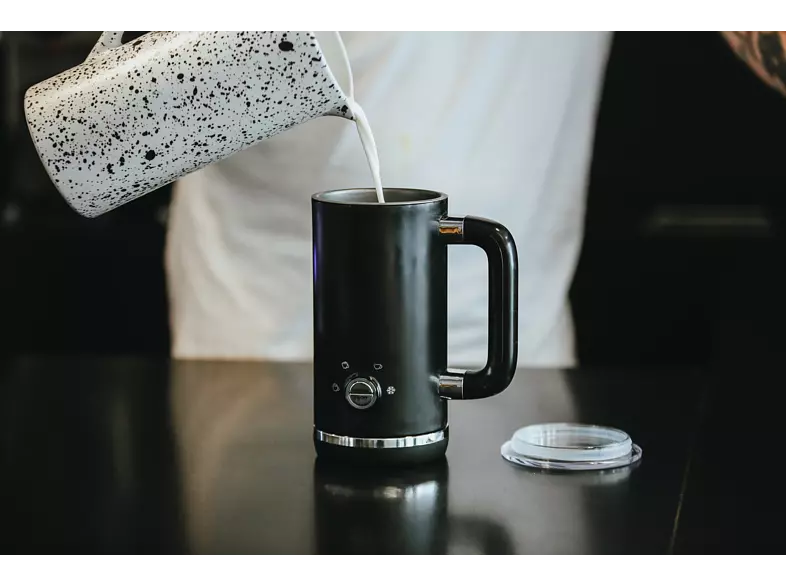 Milk being poured into a black mug with a handle, on a dark surface.