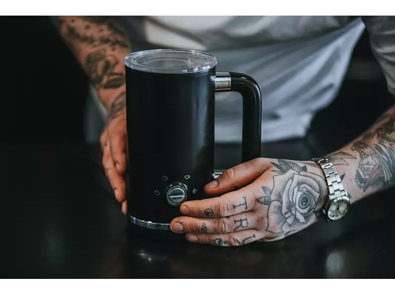A person's tattooed hands hold a black coffee maker with buttons. The handle is black.