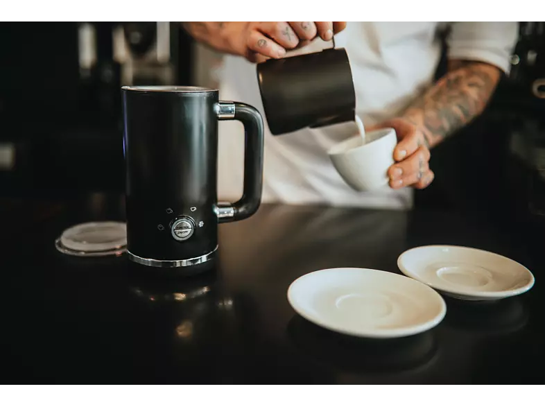 Barista pouring milk into a cup. Black electric milk frother in the background.