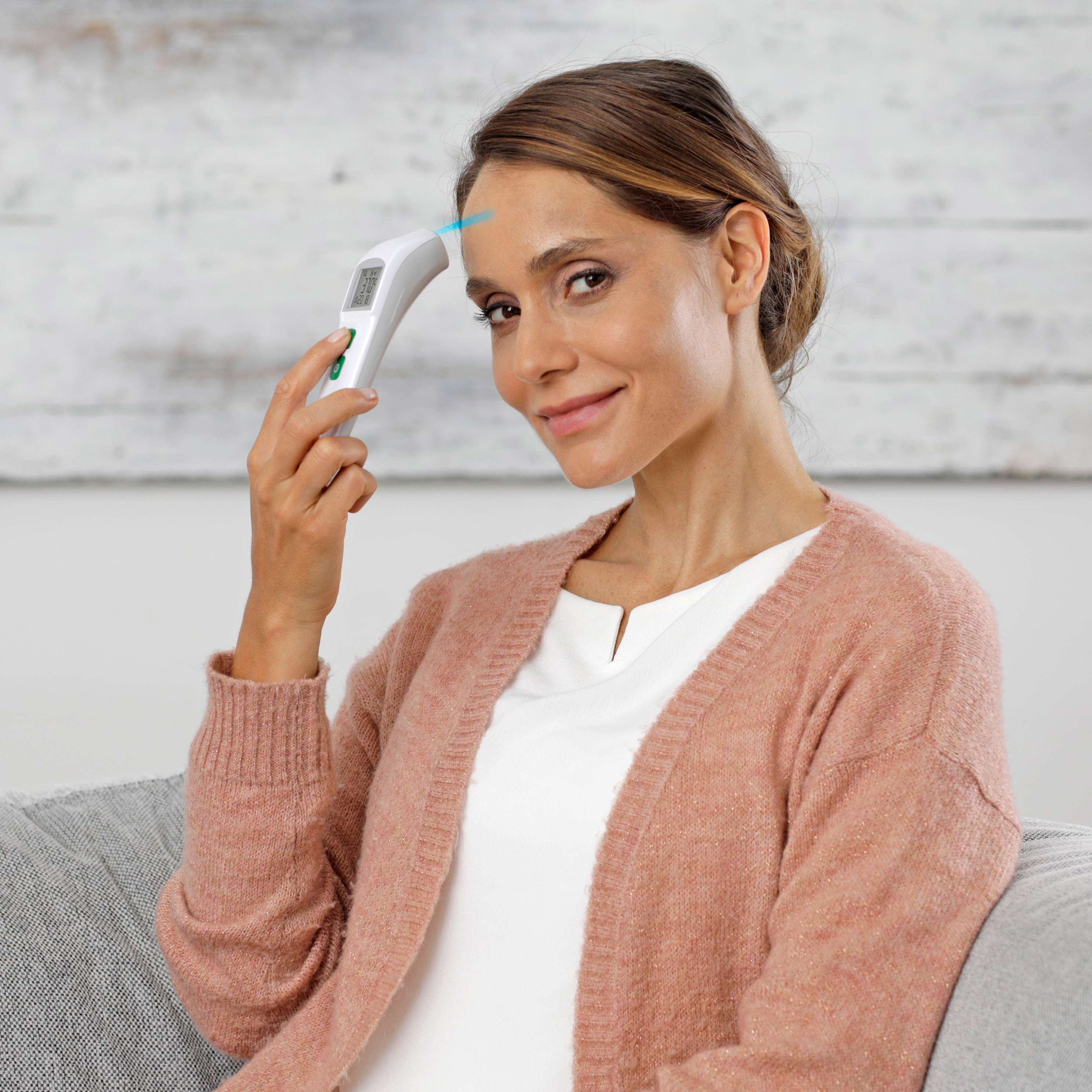 A woman takes her temperature with a non-contact thermometer. The background is a white wall.