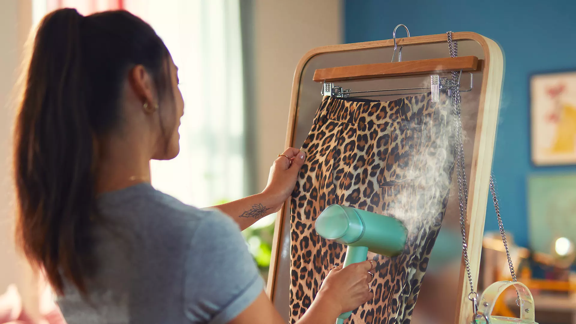 Woman steaming leopard print pants on a mirror. Steamer is light green.