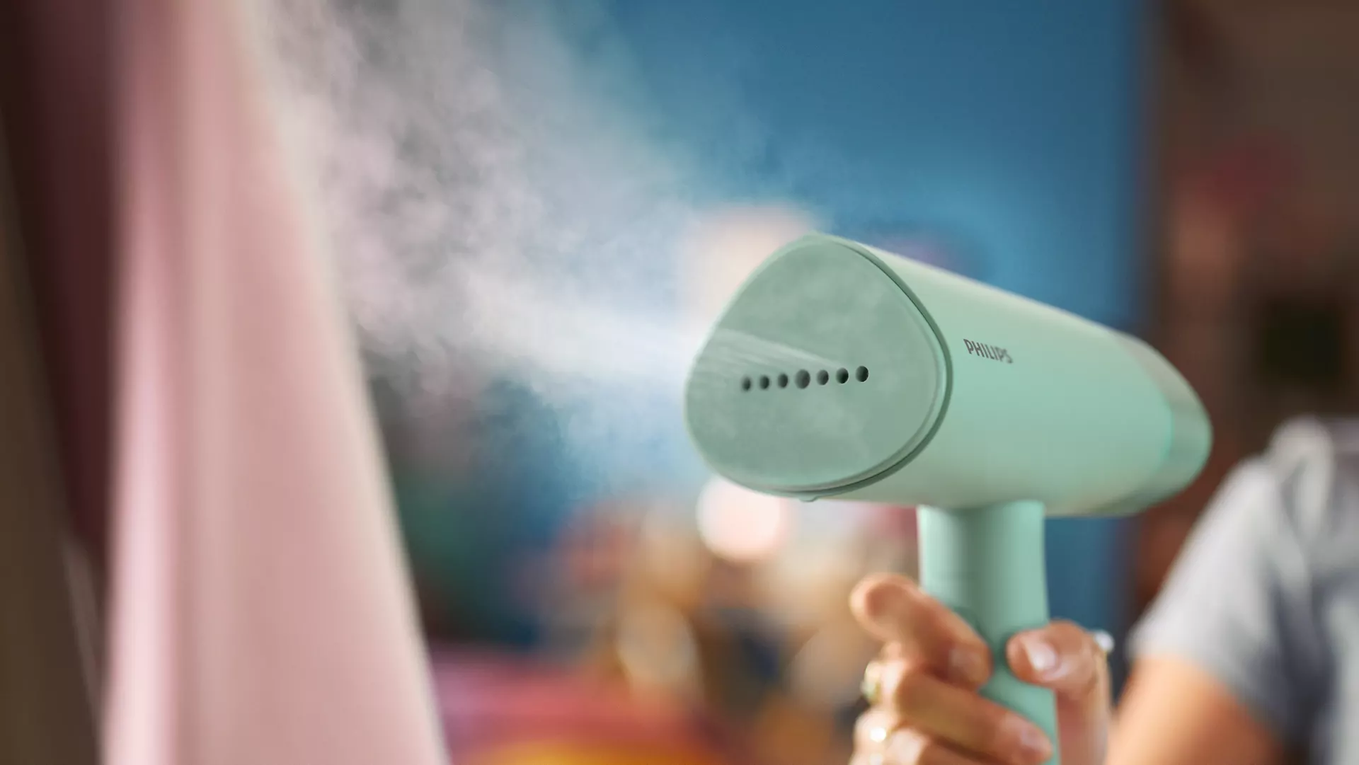 A handheld Philips steamer, mint green, emitting steam over a pink garment. Hand holding steamer.