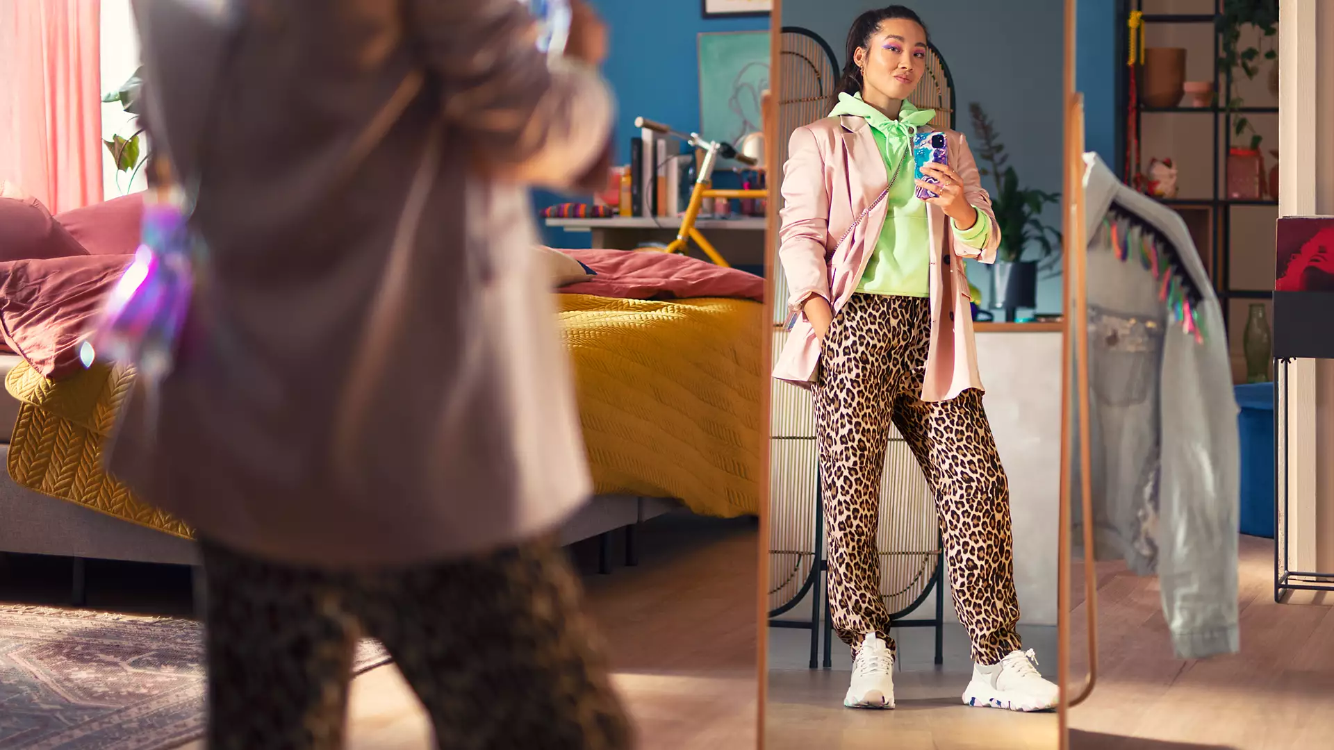 Woman in front of a mirror, wearing leopard print pants, blazer, and a green hoodie, holding a phone.