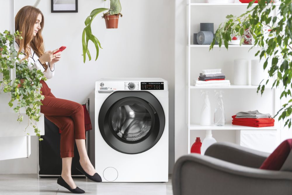 Femme utilisant un téléphone, assise devant un lave-linge blanc, beaucoup de plantes.