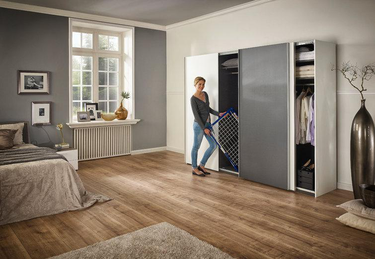 Eine Frau hält ein Wäschegestell vor einem weißen und grauen Kleiderschrank. Ein Bett im Hintergrund.