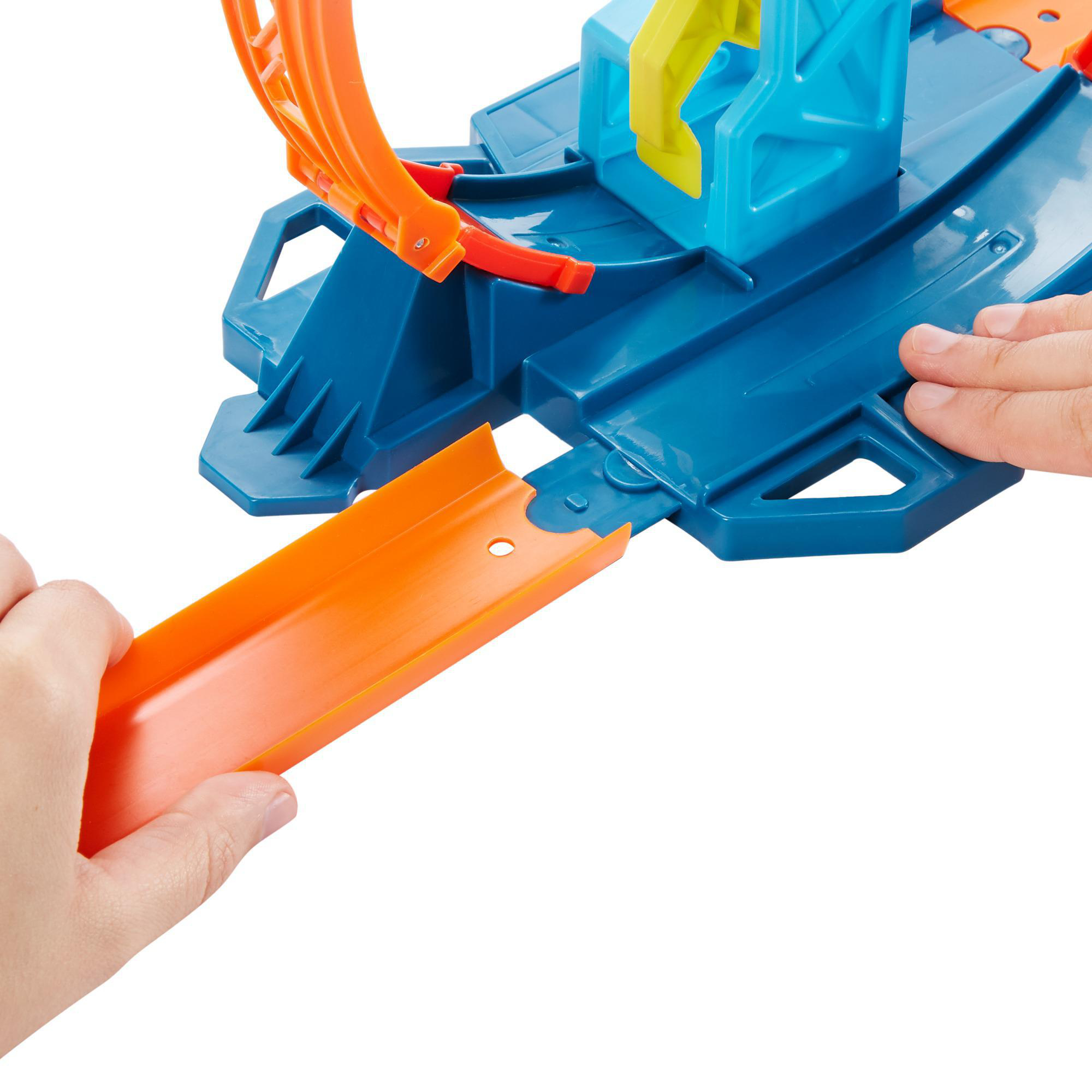 Hände halten eine orangefarbene Spielzeugstrecke, die zu einer blauen Streckenbasis mit gelber Struktur und orangefarbener Schleife führt.