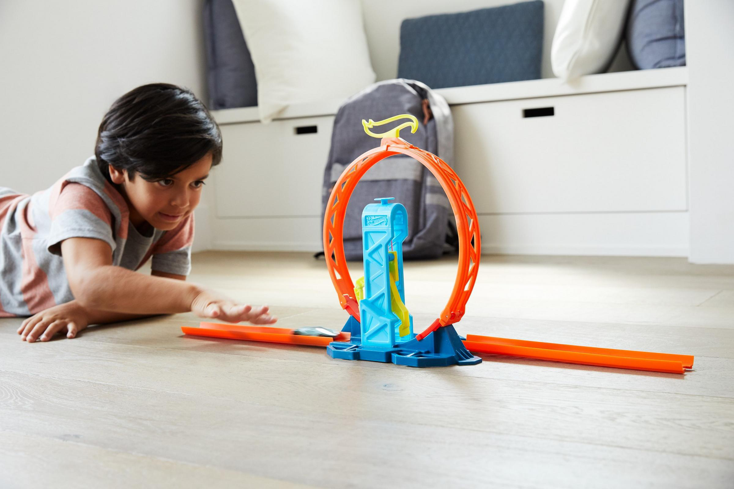 Ein Junge beobachtet eine Spielzeug-Autorennbahn mit einer Loop-de-Loop. Die Strecke ist orange und blau. Ein Rucksack und Kissen sind im Hintergrund.
